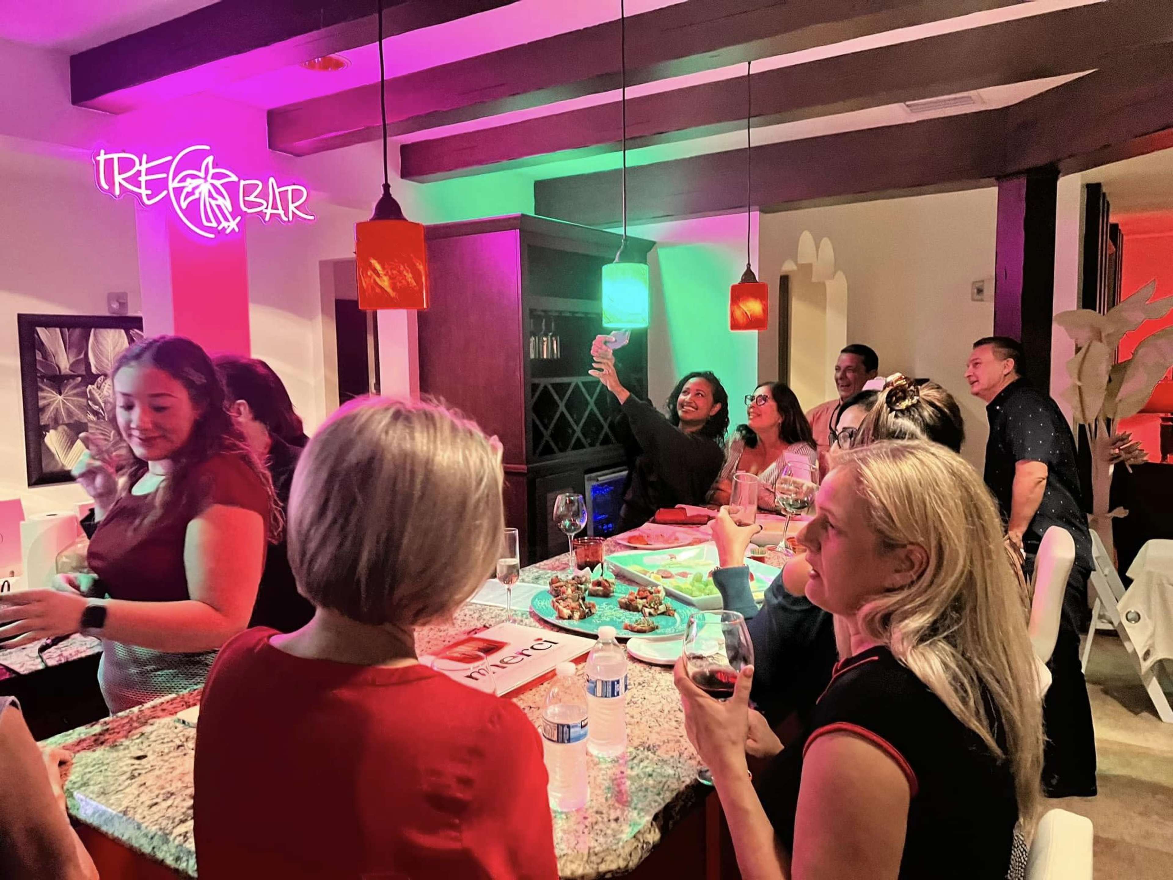 A group of people gathers around a bar area, enjoying drinks and conversation under colorful lights.