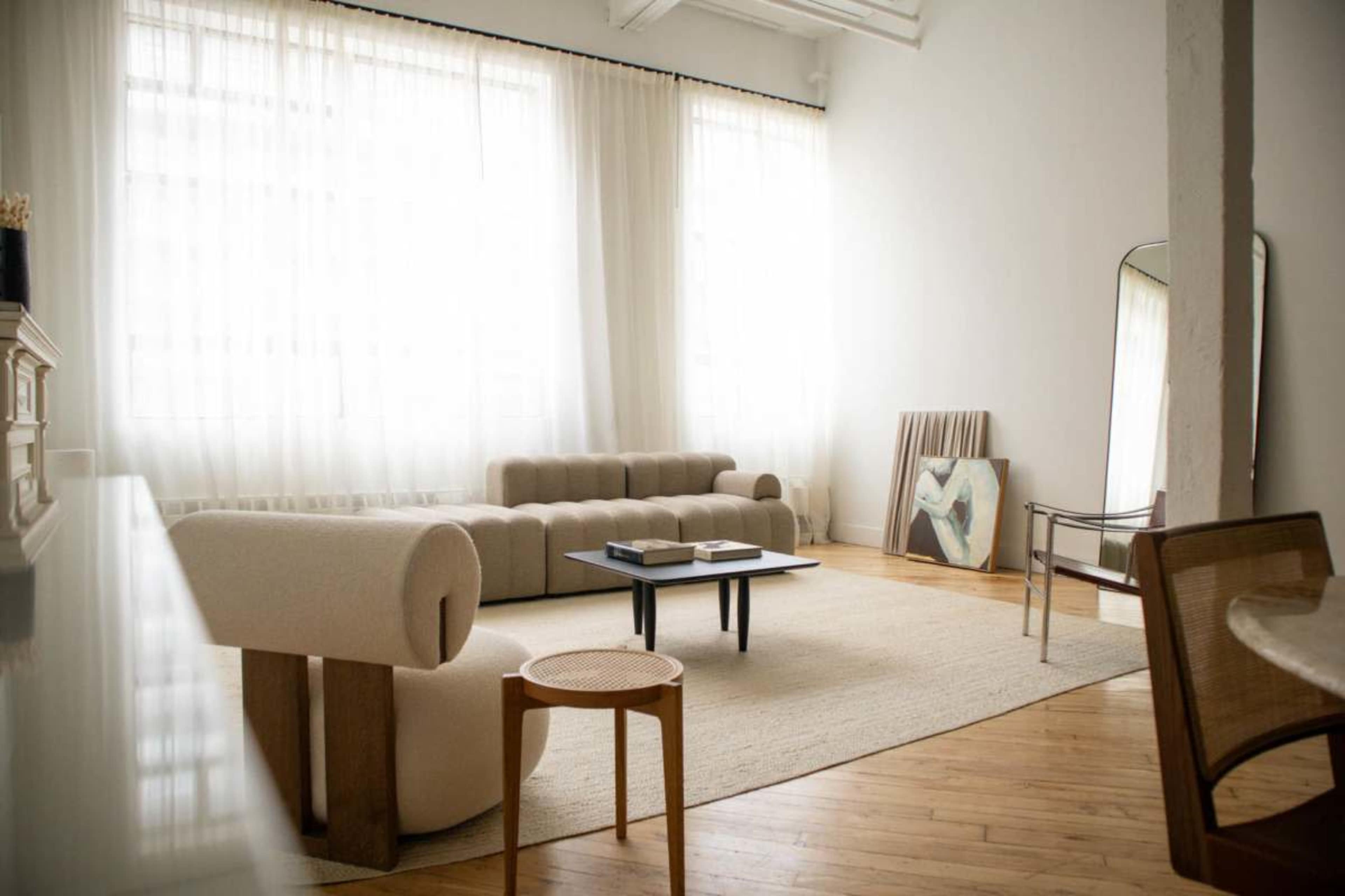 The image depicts a minimalist living room featuring a light-colored rug, two sofas, a coffee table, and a large mirror, with sheer curtains allowing natural light to enter.