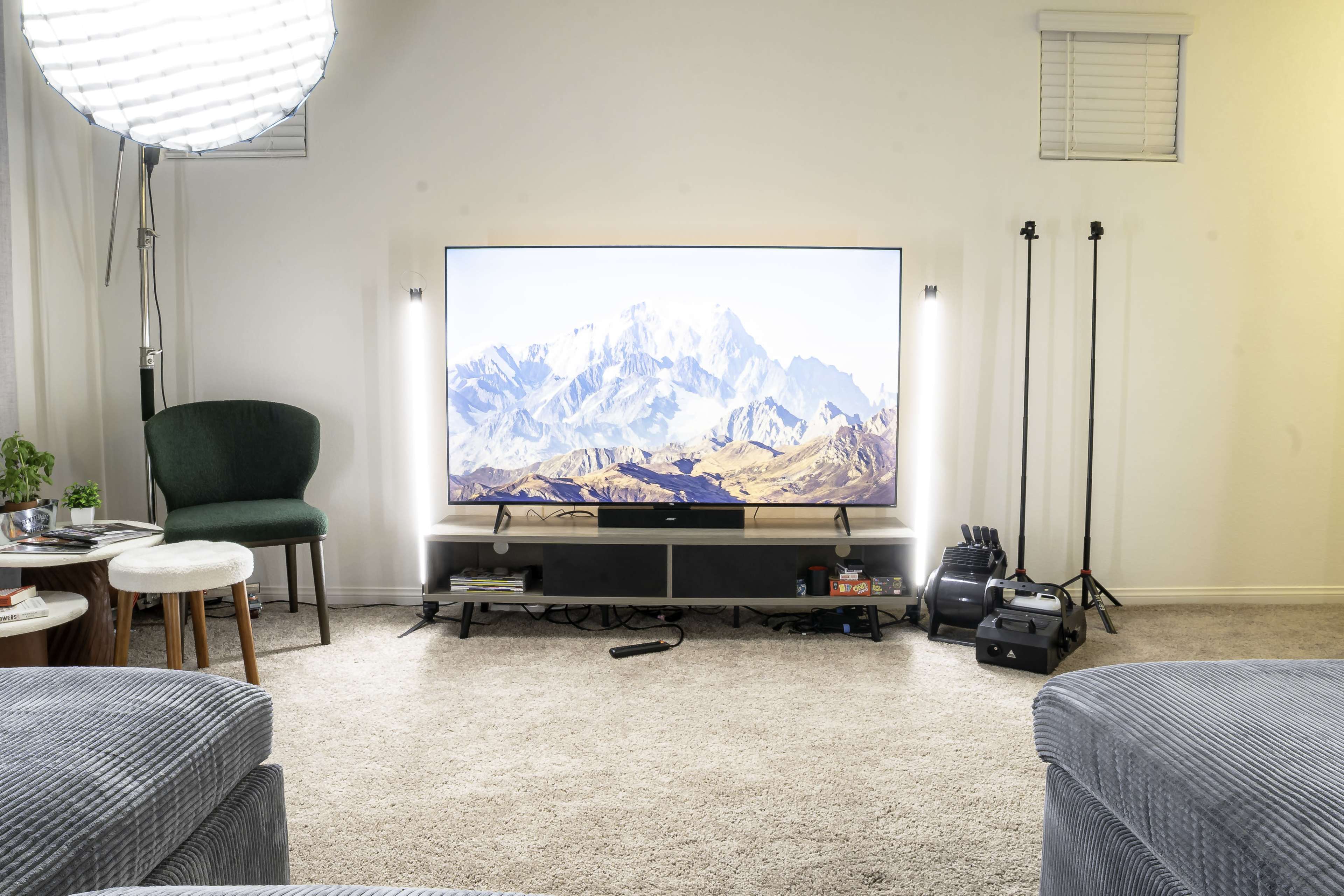 The scene features a living room with a large television displaying a mountain landscape, surrounded by a green chair, a small table, and various equipment including lighting and a projector.