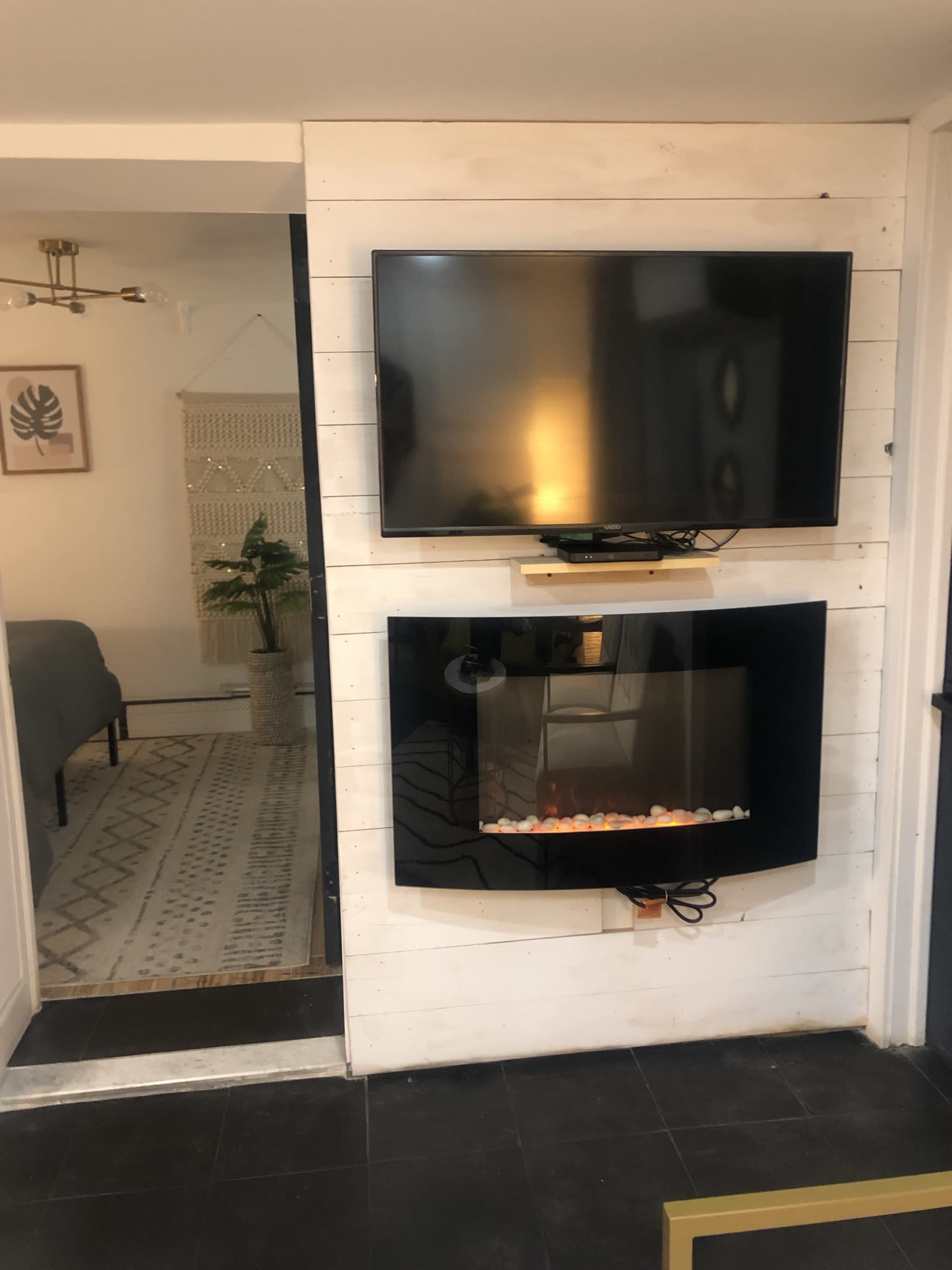 The image shows a living space with a mounted flat-screen TV above a modern electric fireplace, both set against a white wooden wall.