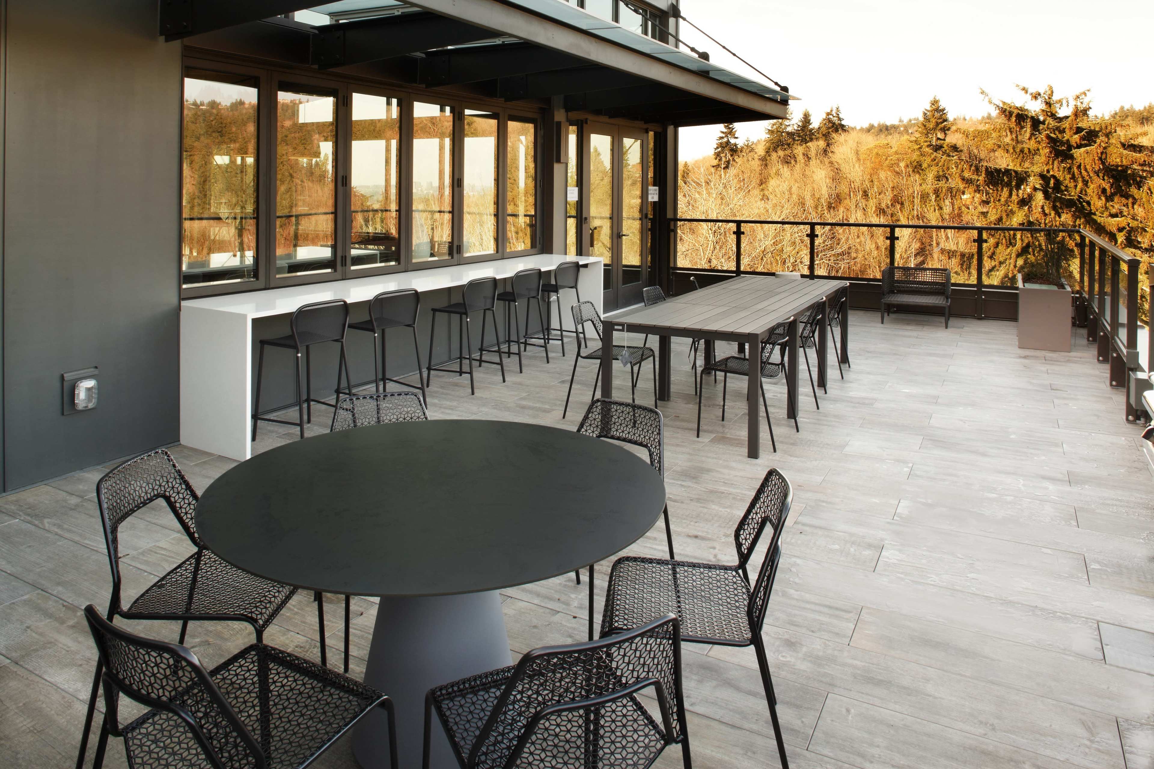 A modern outdoor terrace with a circular table and several chairs, along with a long rectangular table and additional seating, set against a backdrop of trees and a clear sky.