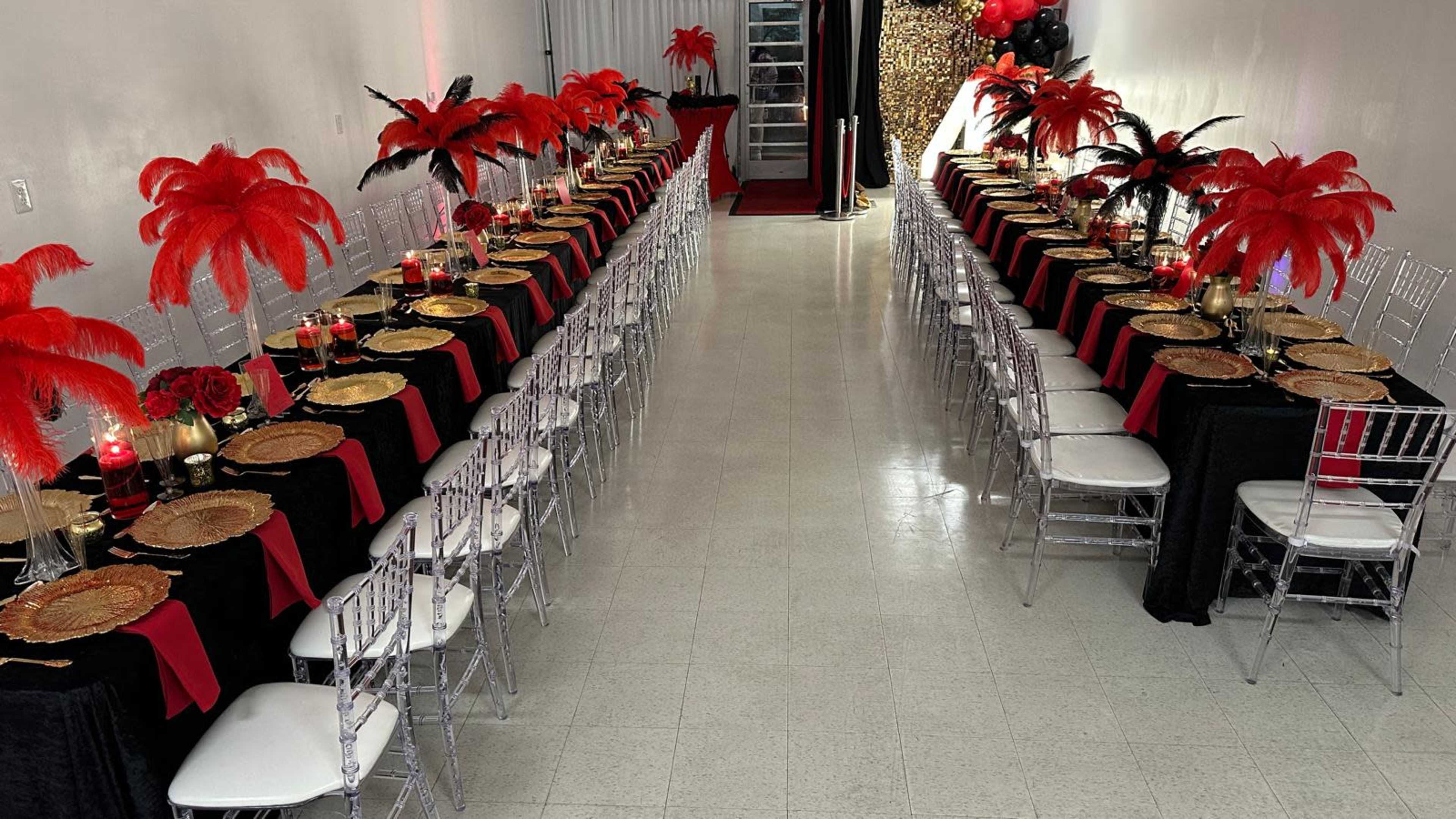 The image shows a banquet hall set with two long tables covered in black and red tablecloths, adorned with golden plates and red feather centerpieces.