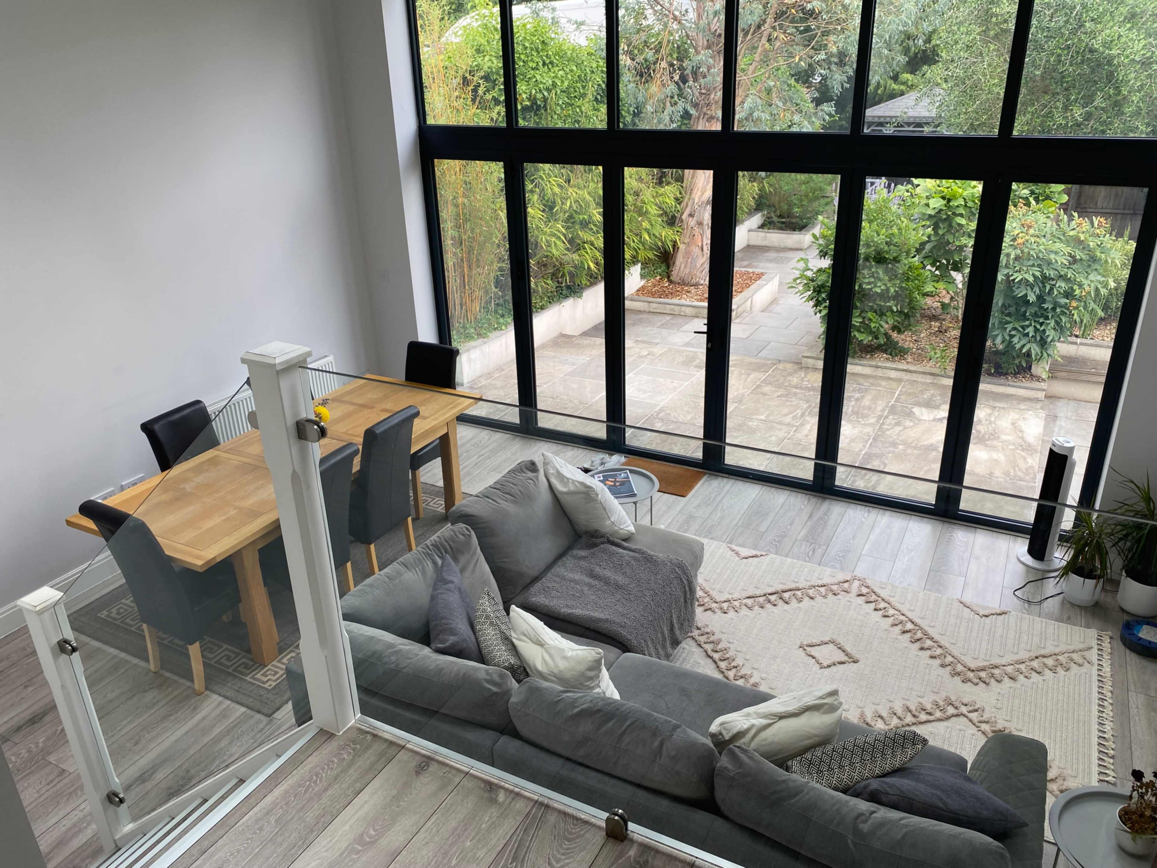 A modern living space features a gray sectional sofa, a wooden dining table with chairs, and large glass doors overlooking a garden.