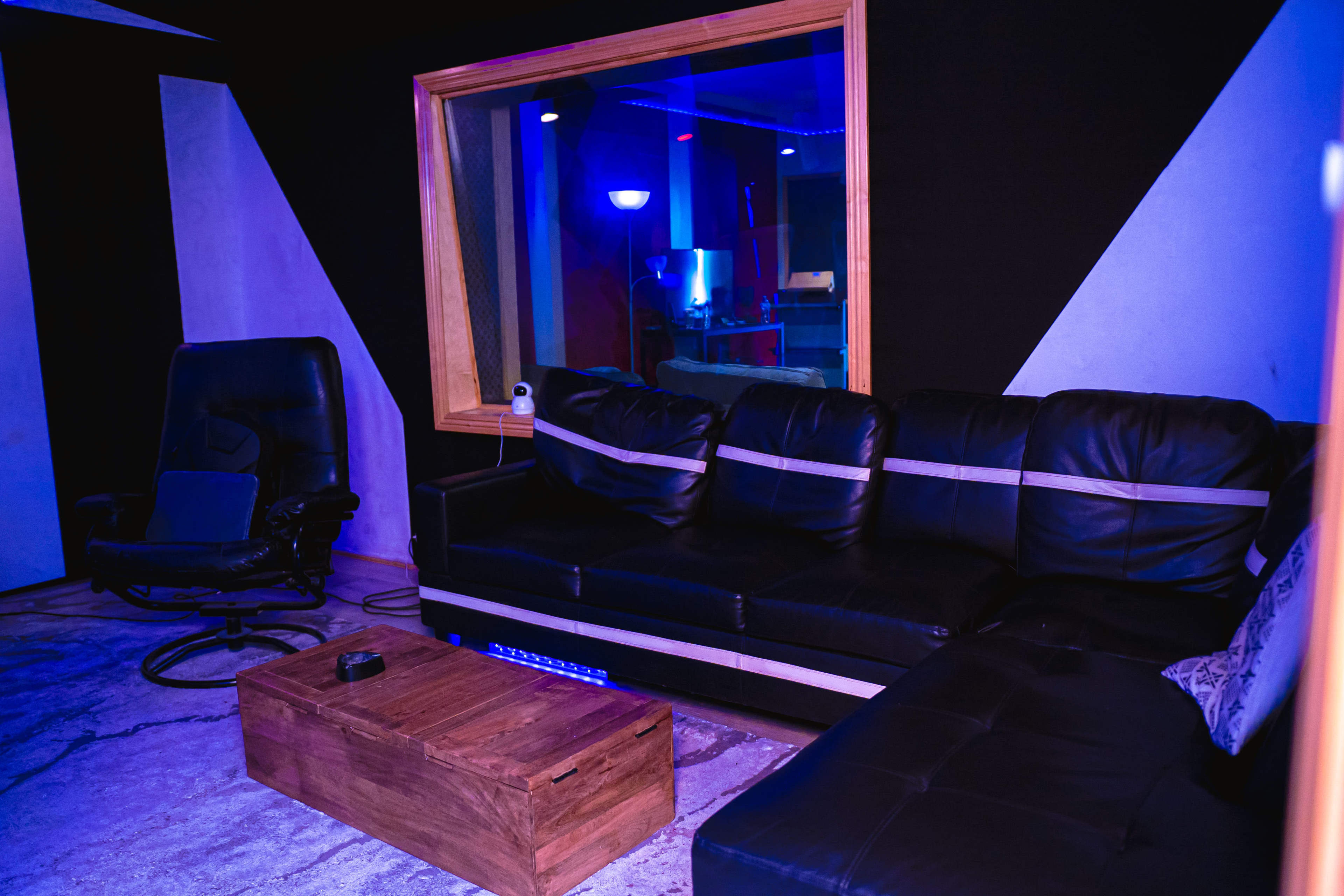 A dimly lit recording studio with a black leather couch, a wooden coffee table, and a cozy recliner chair.