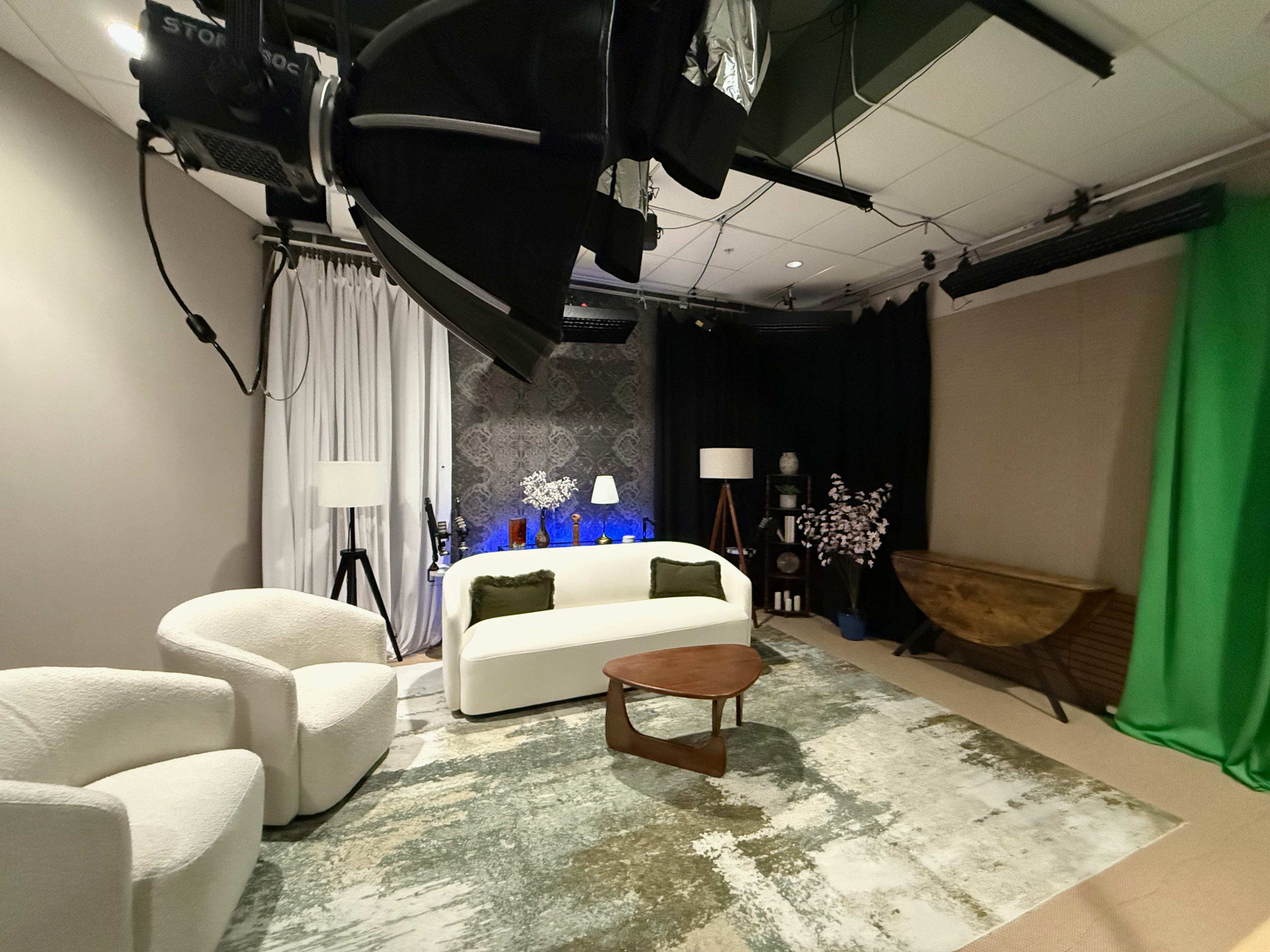 The image shows a cozy studio setting with a white sofa and chairs on a patterned rug, accompanied by a decorative table, lamps, and a green screen backdrop.
