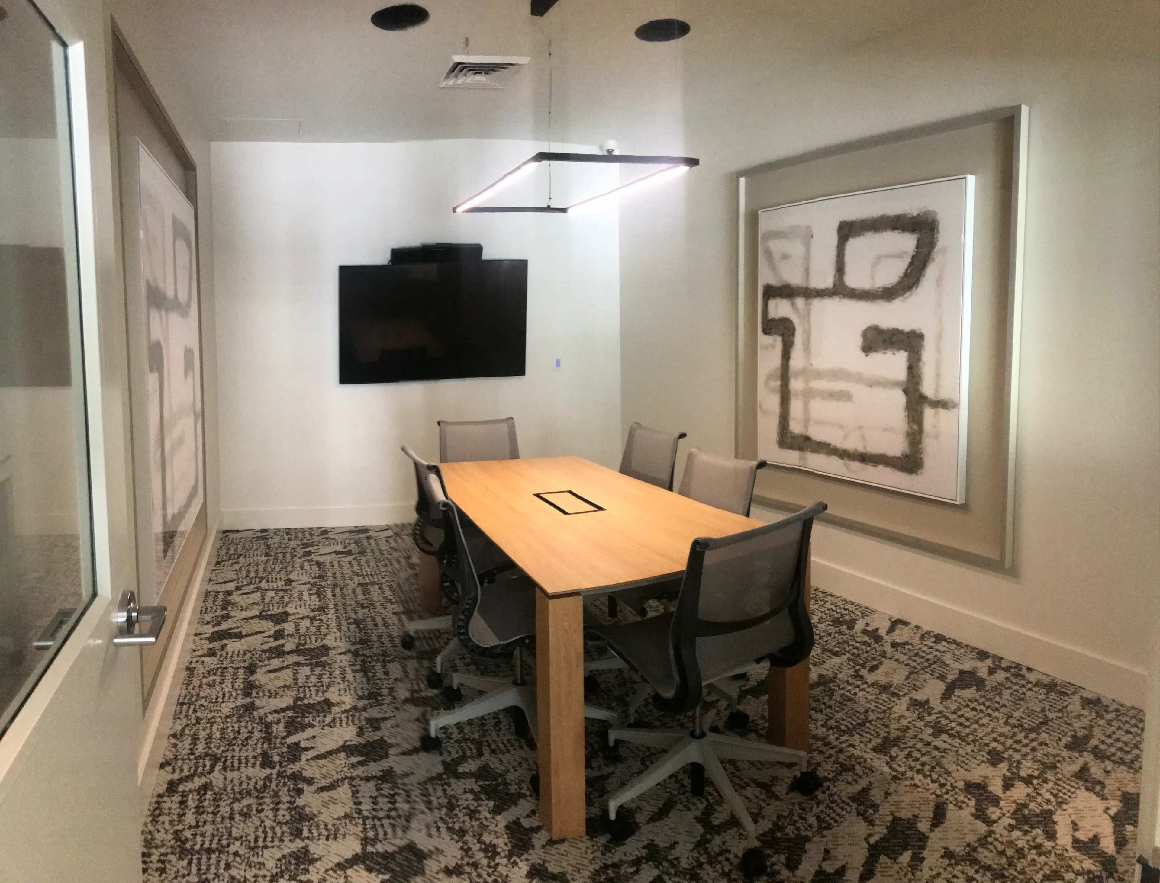 A modern conference room with a wooden table surrounded by four chairs, a large wall-mounted screen, and abstract art on the wall.
