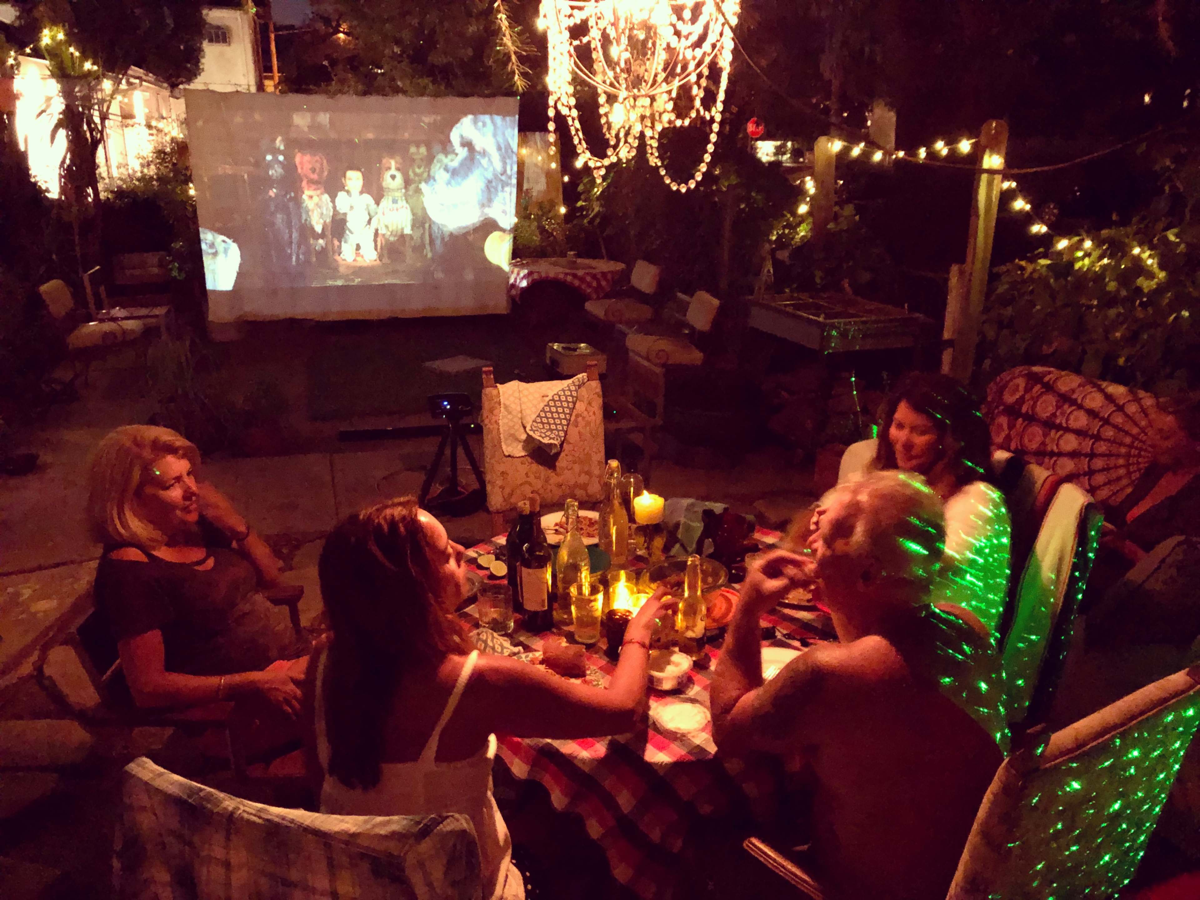 A group of five people sits around a table in a dimly lit outdoor setting, enjoying food and drinks while a movie is projected on a nearby wall.
