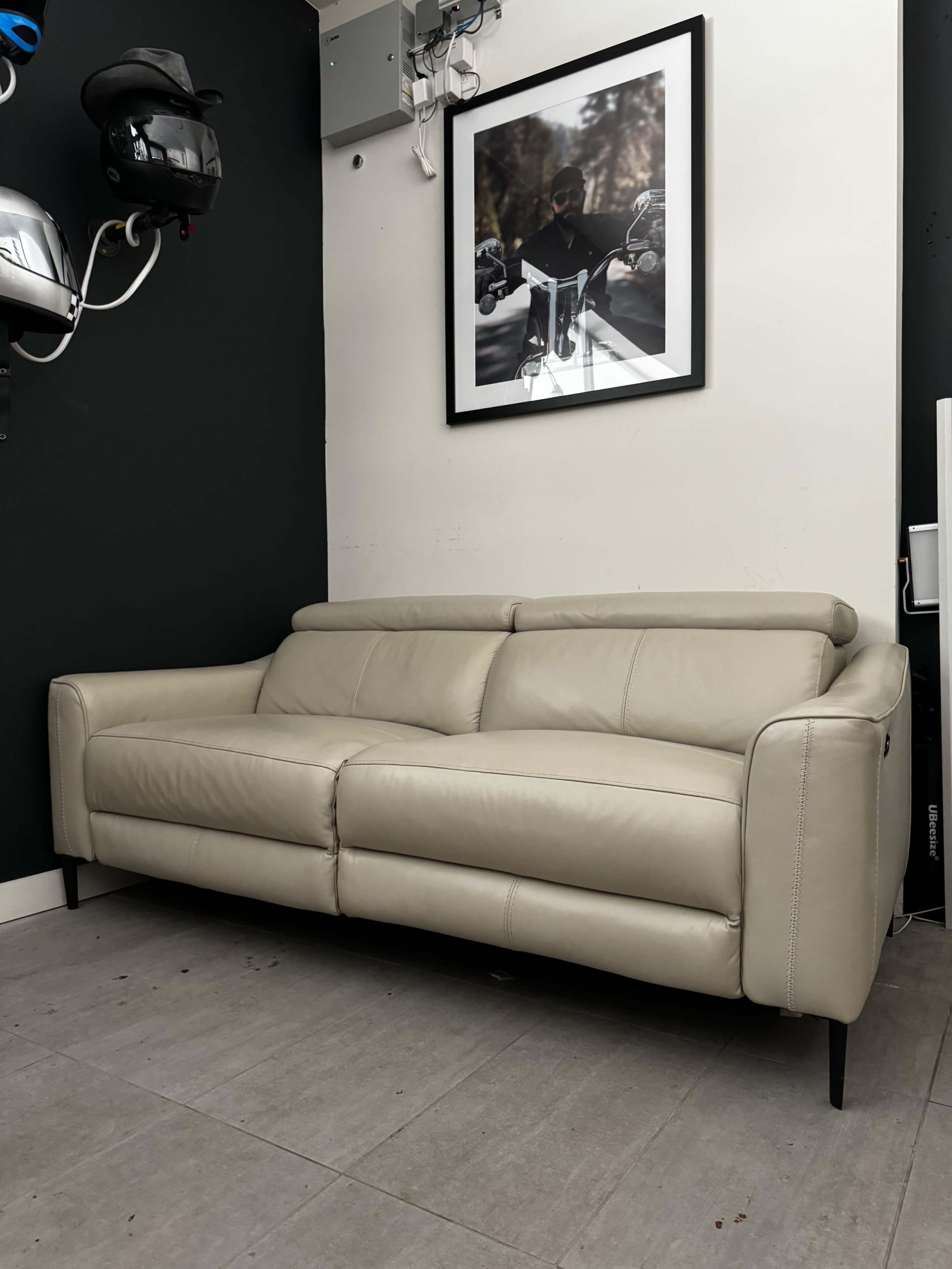 A light-colored leather sofa positioned against a dark wall, next to a framed photograph and motorcycle helmets hanging on the side.