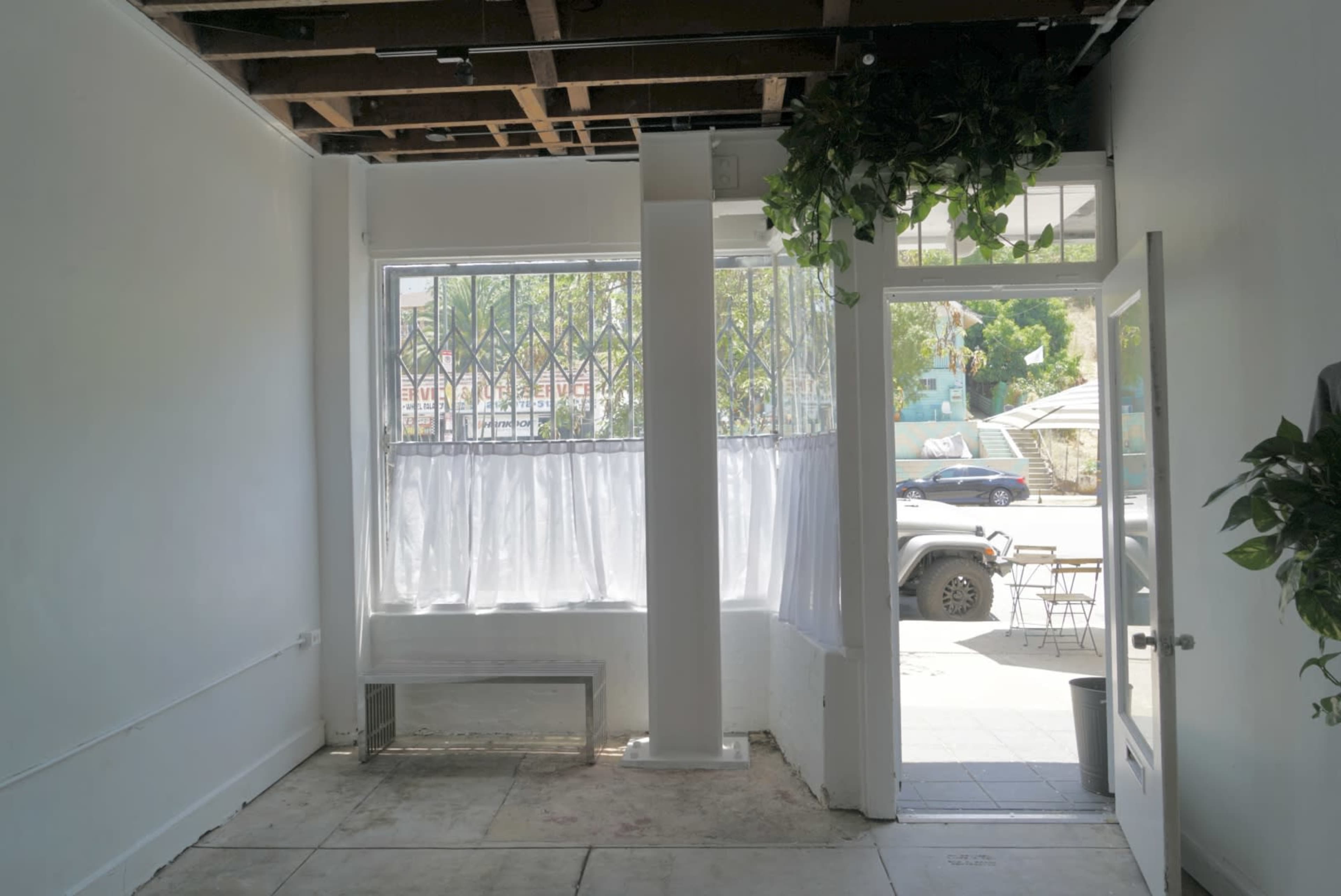 A sparse interior space with white walls, a large window covered by white curtains, and an open door leading outside.