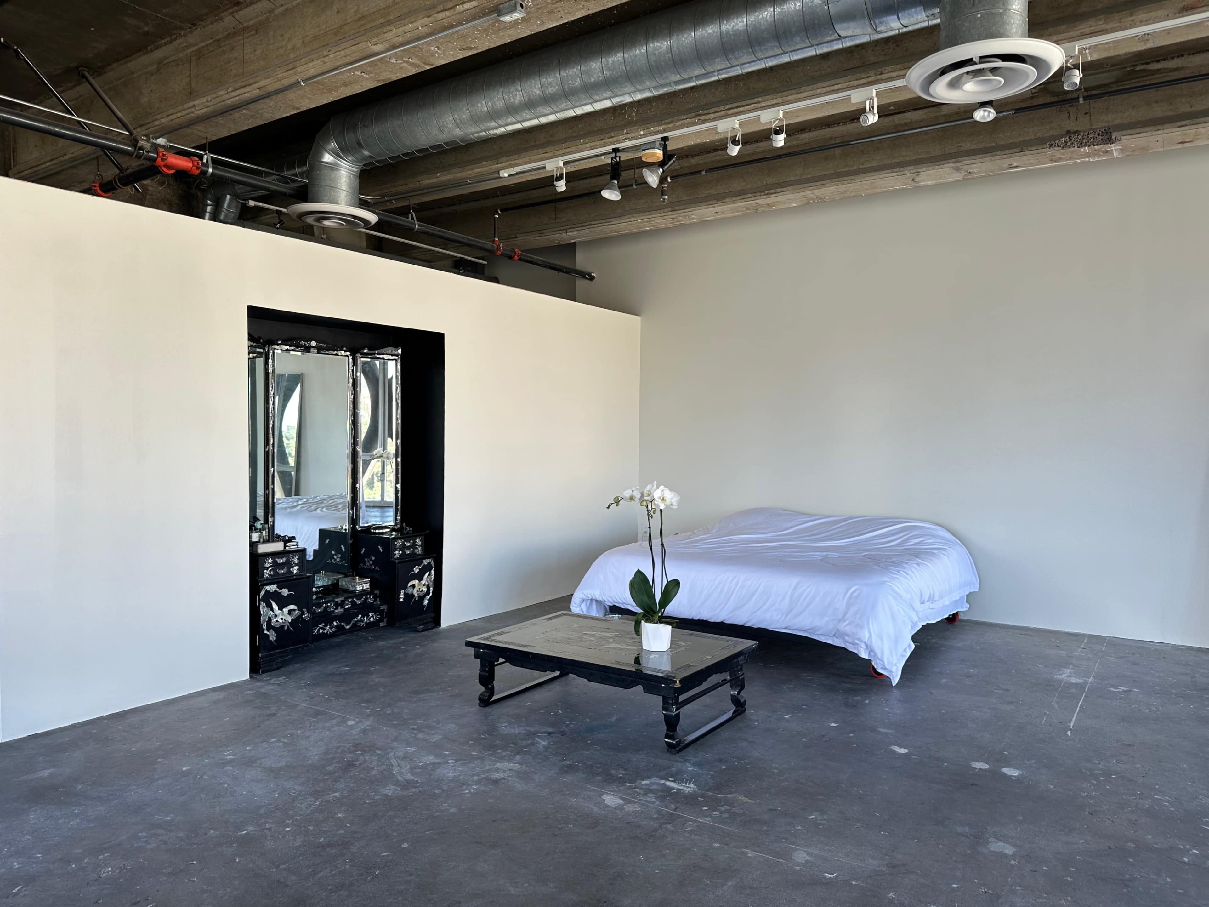 The image shows a minimalist interior with a bed covered in a white sheet, a black table with a plant, and a mirrored wall in an industrial-style space.