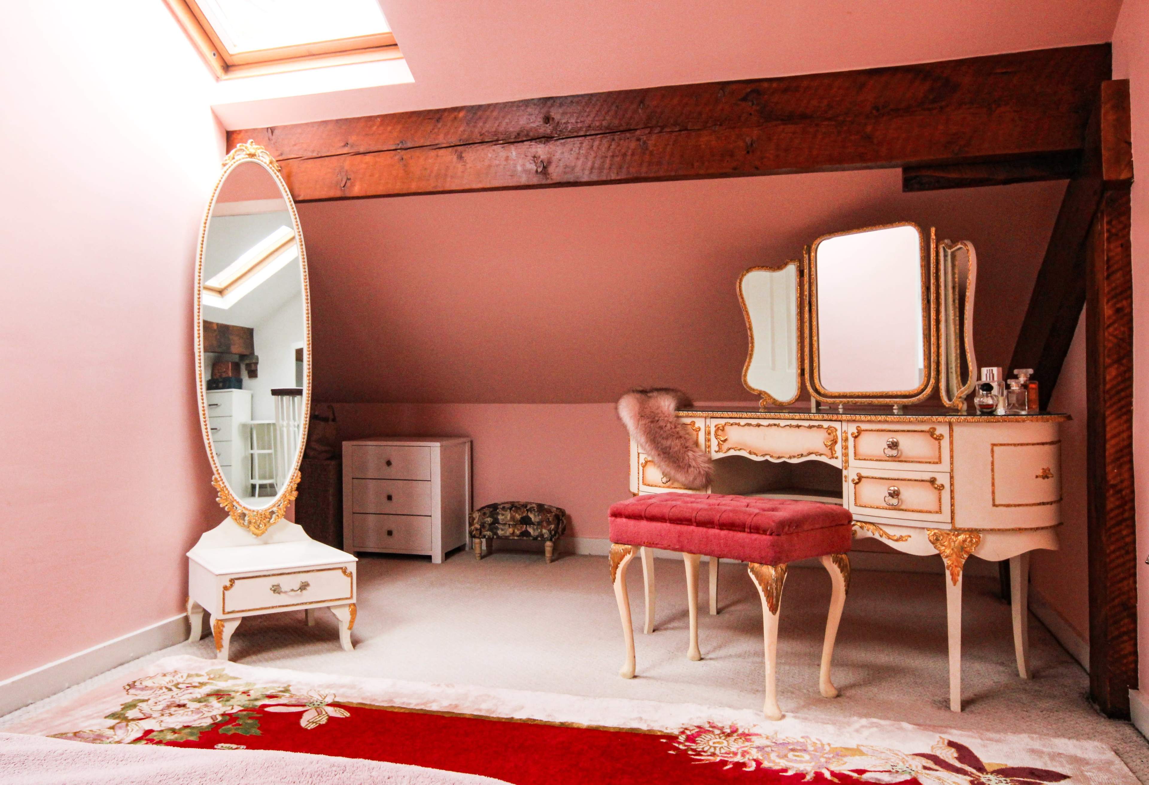 A softly lit bedroom features a vintage vanity with a pink cushioned stool, a large ornate mirror, and a small dresser against a pale pink wall with exposed wooden beams.
