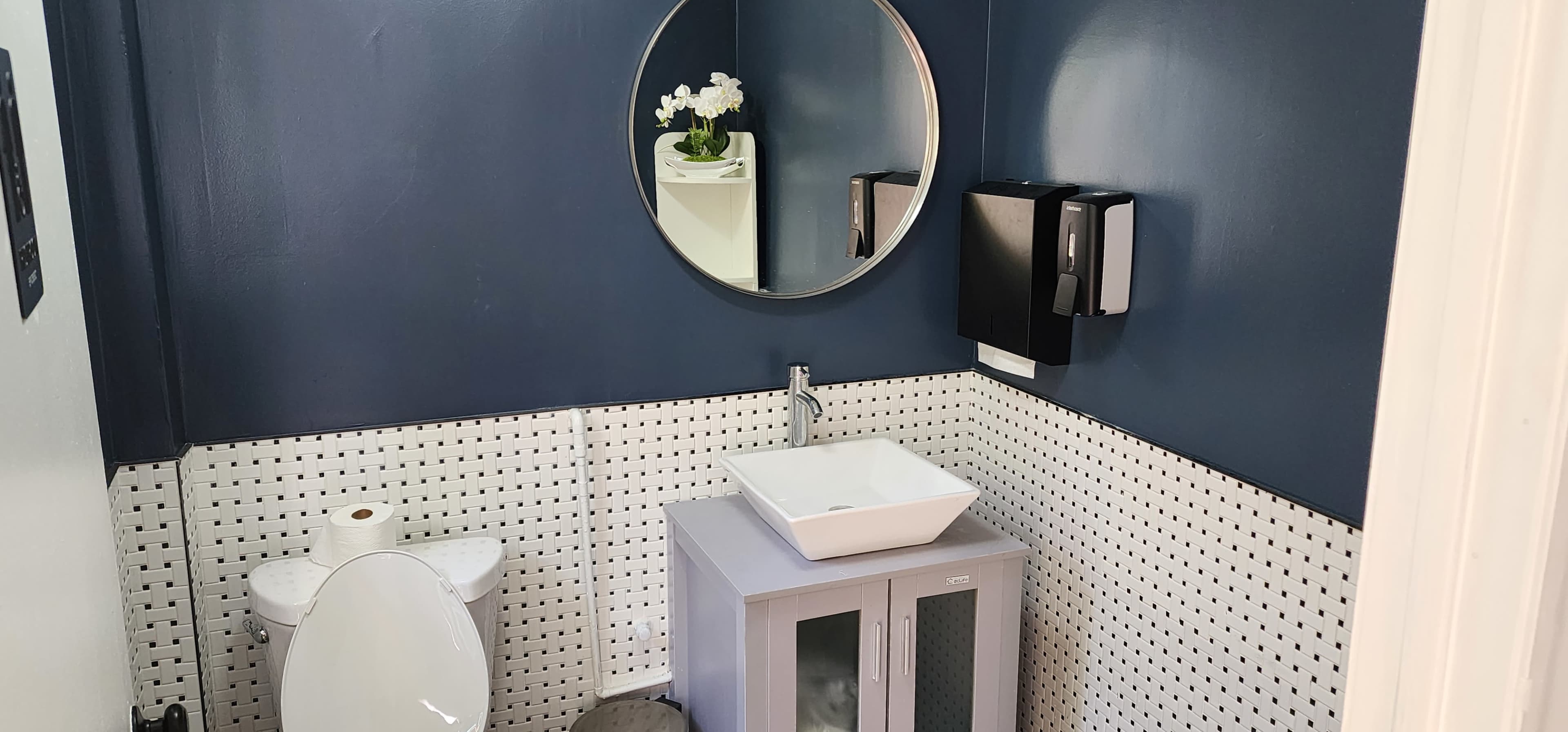 A modern bathroom with a navy-blue wall, a circular mirror, a white sink on a gray cabinet, a toilet, and a paper towel dispenser.