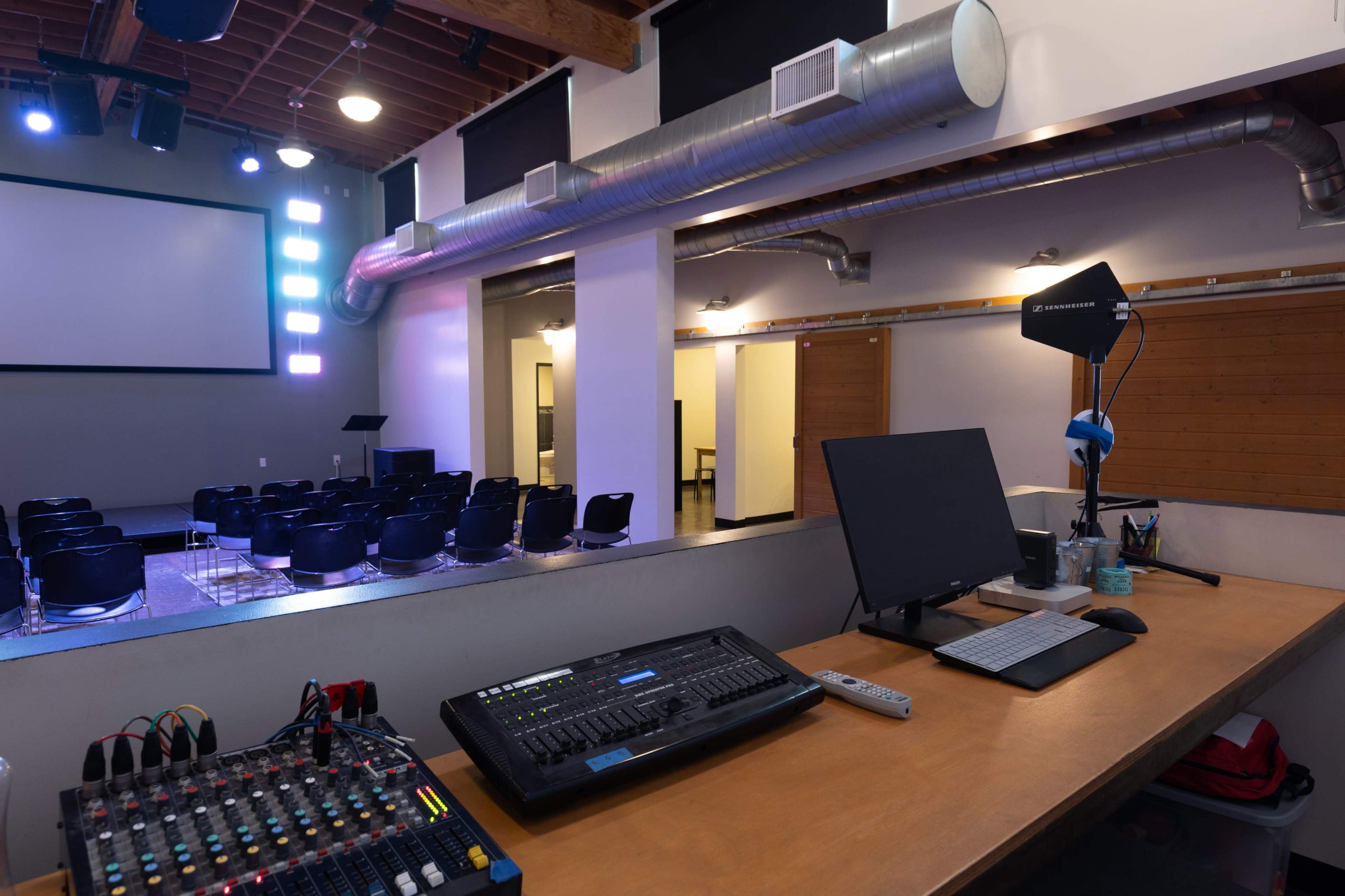 The image shows an interior setting with a control desk featuring a mixing console and a computer, overlooking a stage area with rows of black chairs and a large screen.