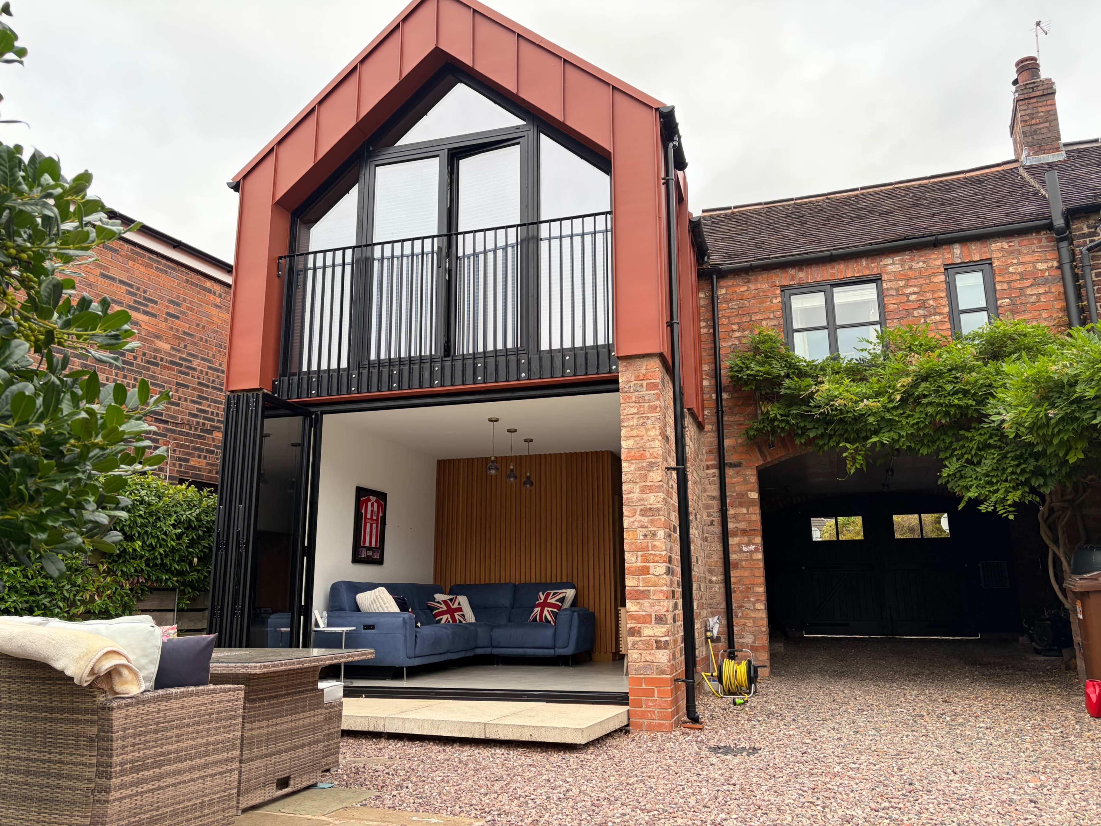 The image shows a modern house with a red metal roof and large glass doors opening to a stylish living area, surrounded by brick walls and a gravel driveway.