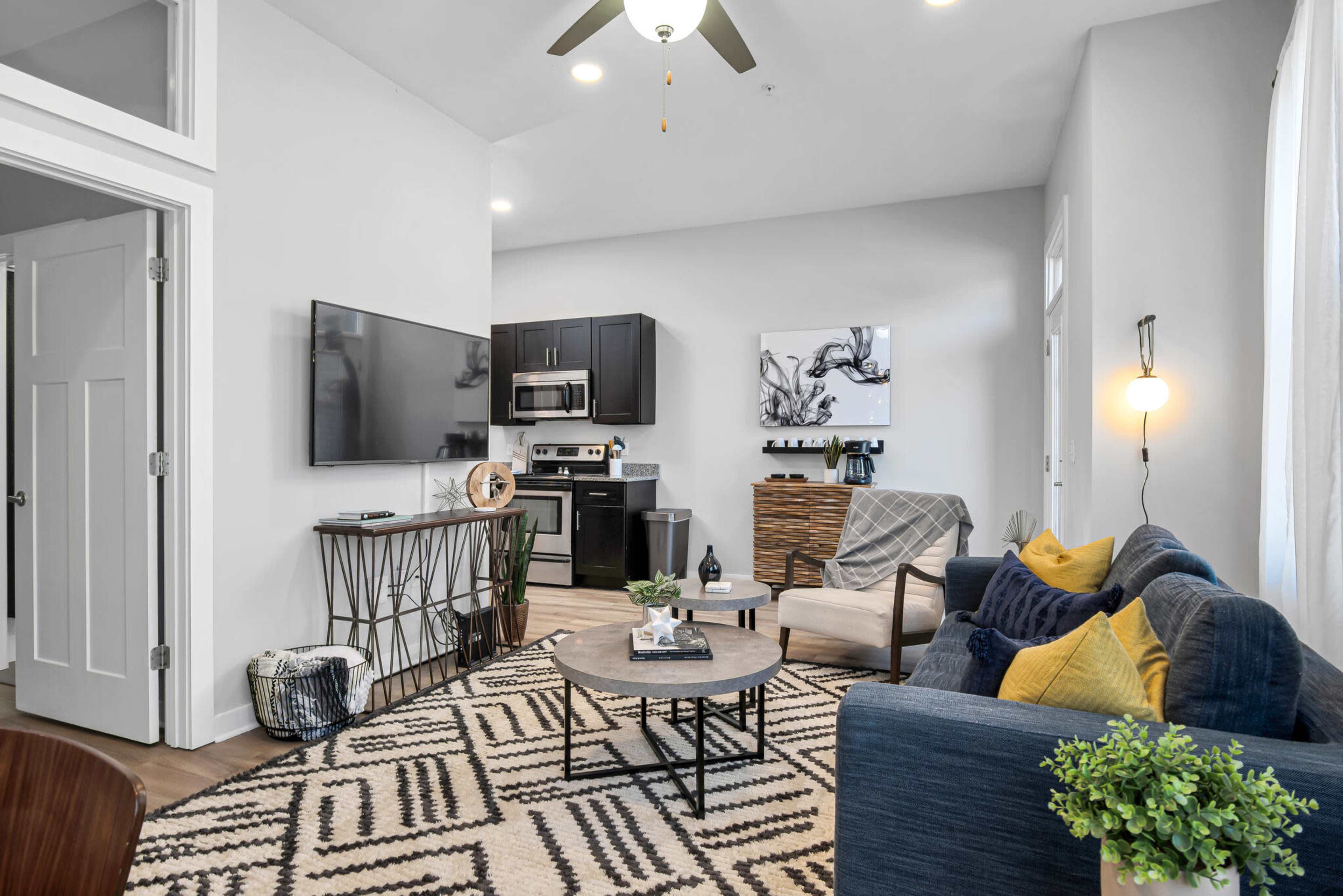 The image shows a modern living space featuring a kitchen area with dark cabinetry, a television mounted on the wall, a round coffee table, and seating arrangements that include a blue sofa and a patterned rug.
