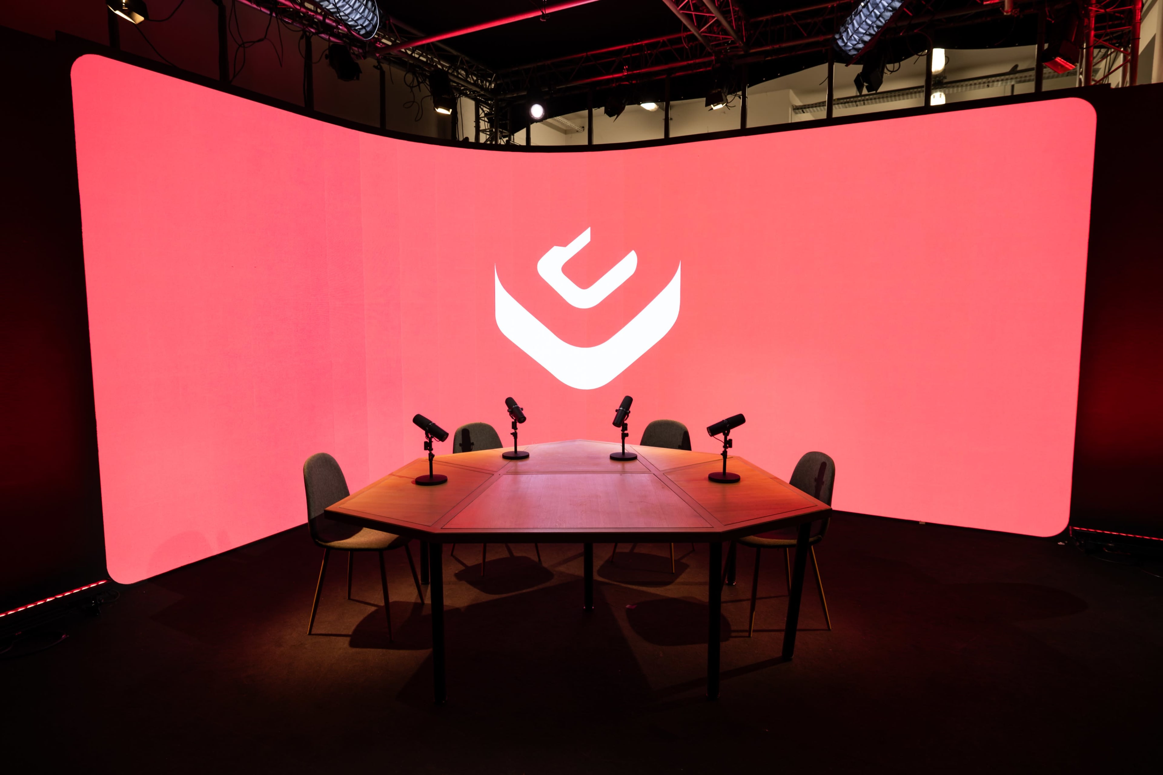 A circular wooden table with four chairs and microphones is set in front of a large red backdrop featuring a white logo.