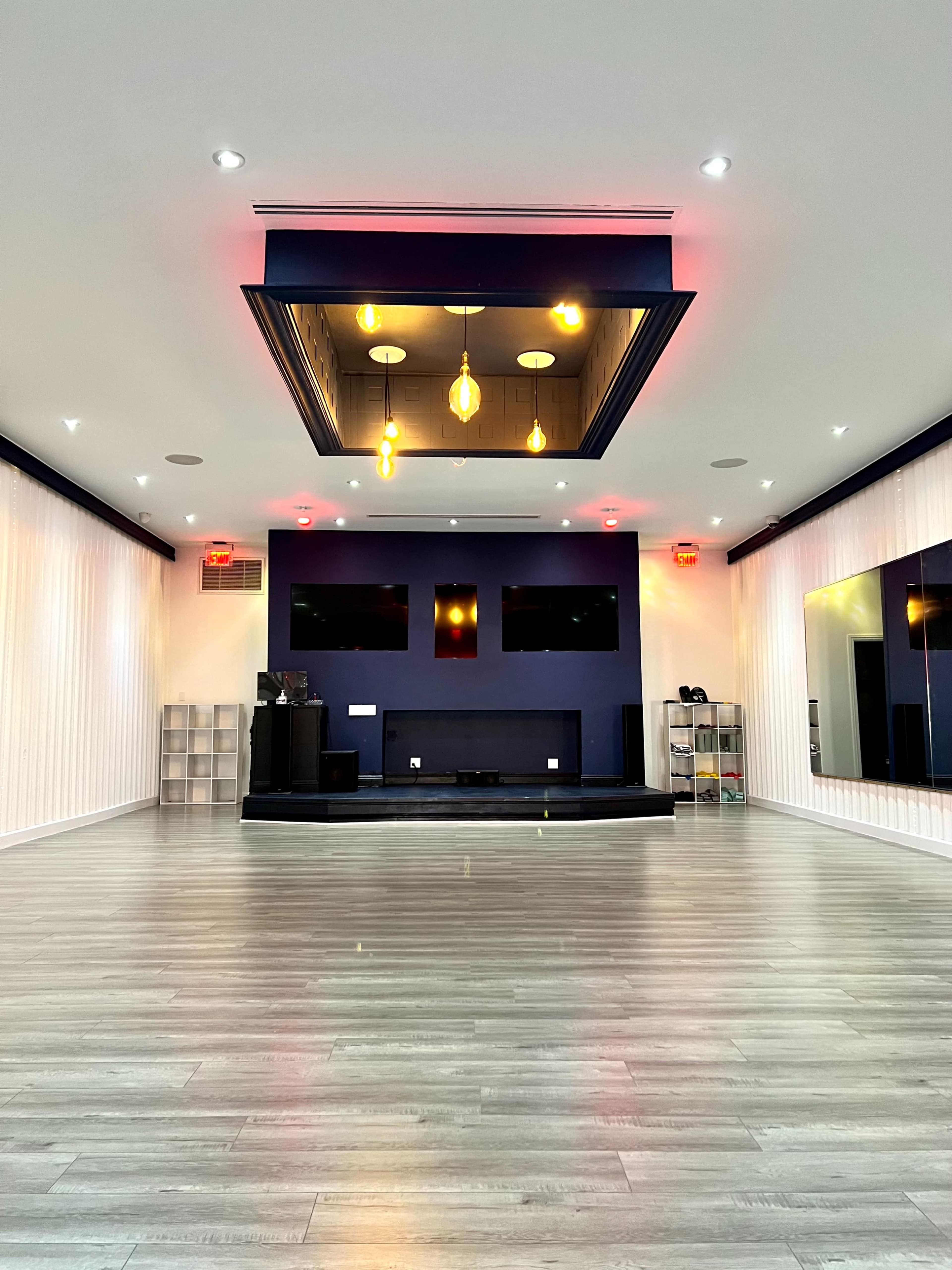 The image shows a modern, empty interior space with wooden flooring, a raised stage, and decorative lighting fixtures on a ceiling feature.
