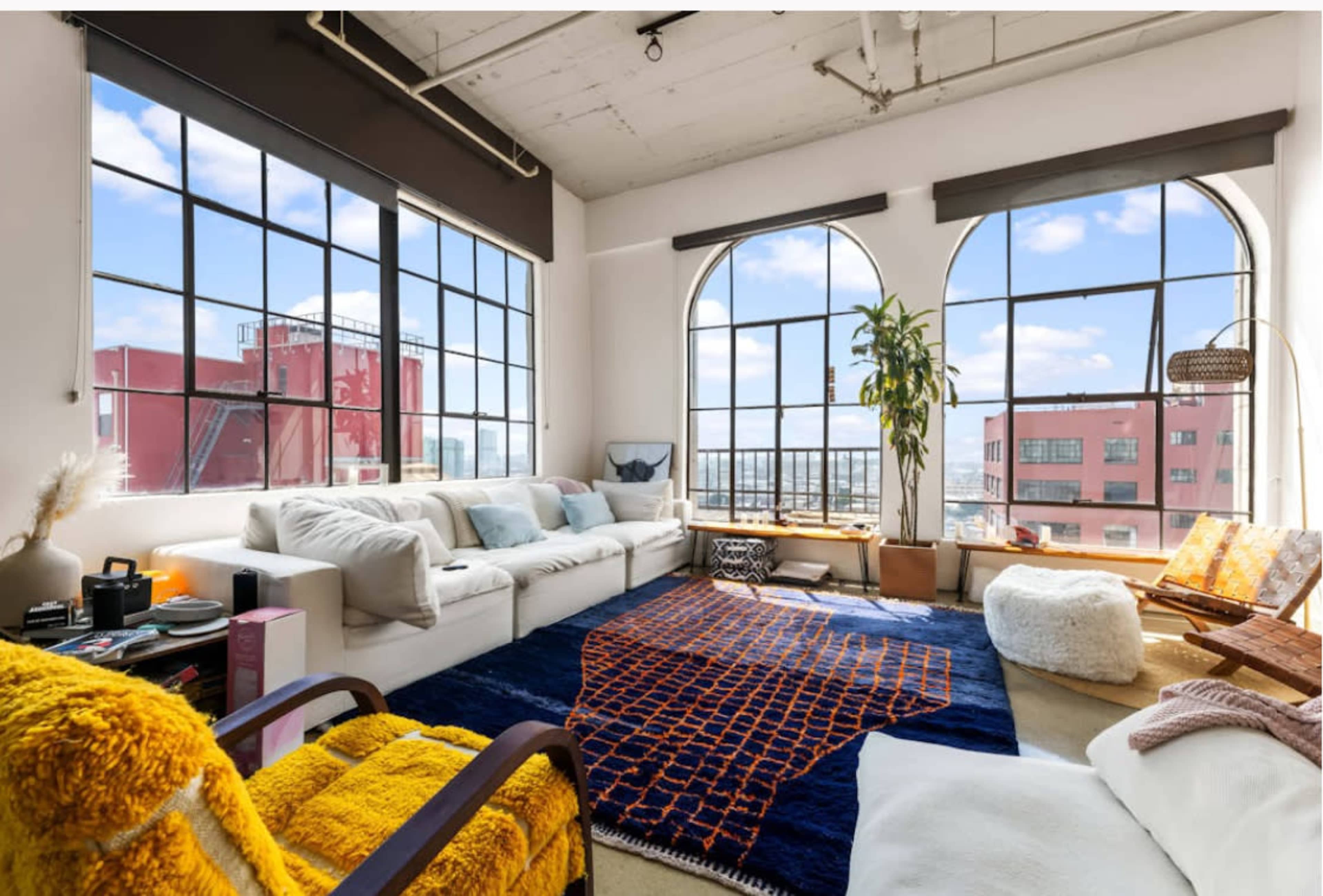 The image shows a bright, modern living room with large arched windows, a cozy sofa, a vibrant area rug, and a tall indoor plant.