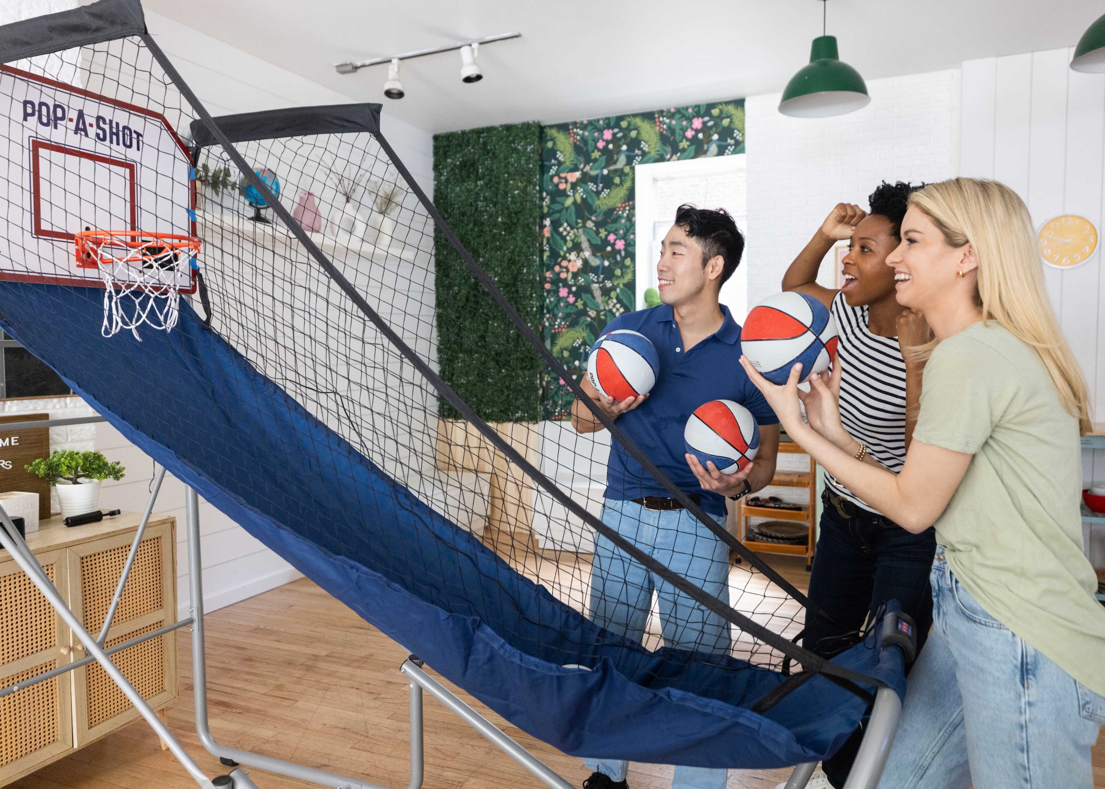 Three people are playing a basketball arcade game inside a well-lit room with indoor plants.