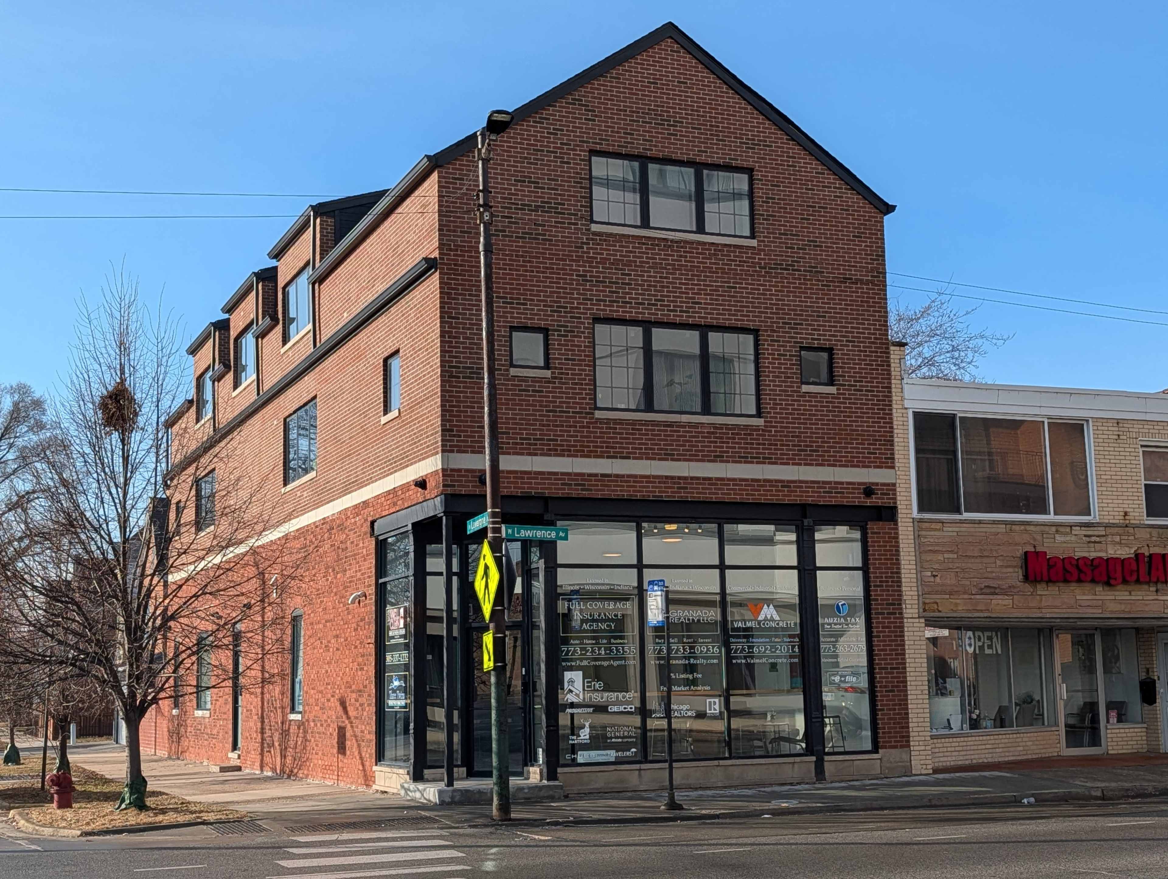The image shows a modern brick building at the corner of a street, featuring large glass windows and multiple stories.