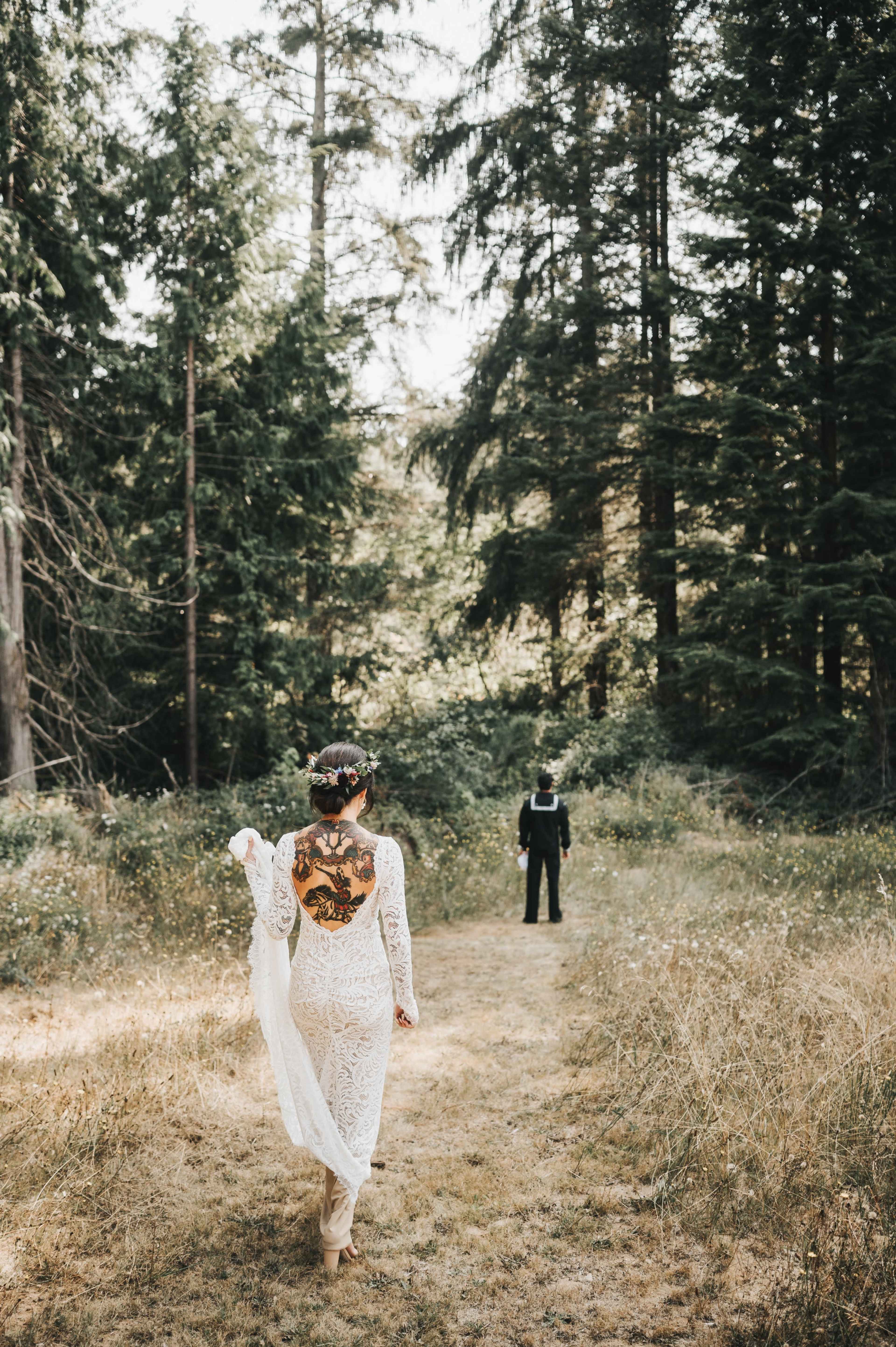 A bride in a lace dress walks down a forest path toward a groom in a dark suit.