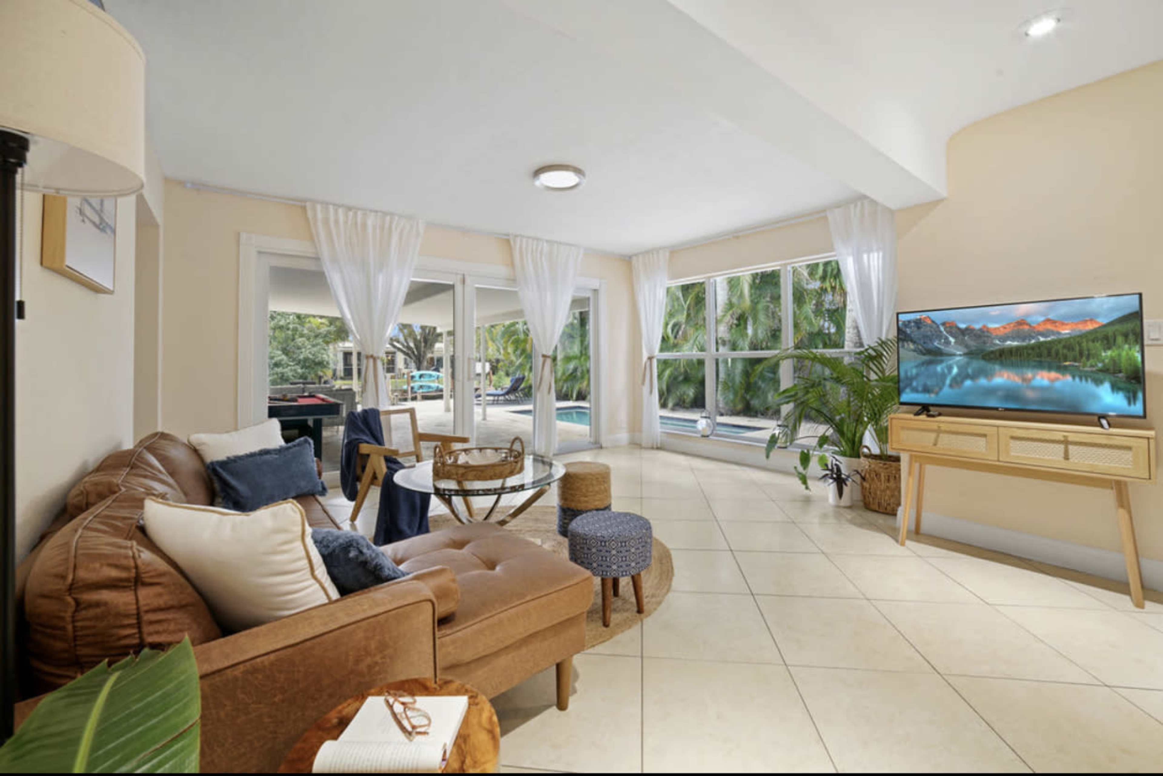 A well-lit living room with large windows, a brown sofa, a coffee table, and a television displaying a nature scene.