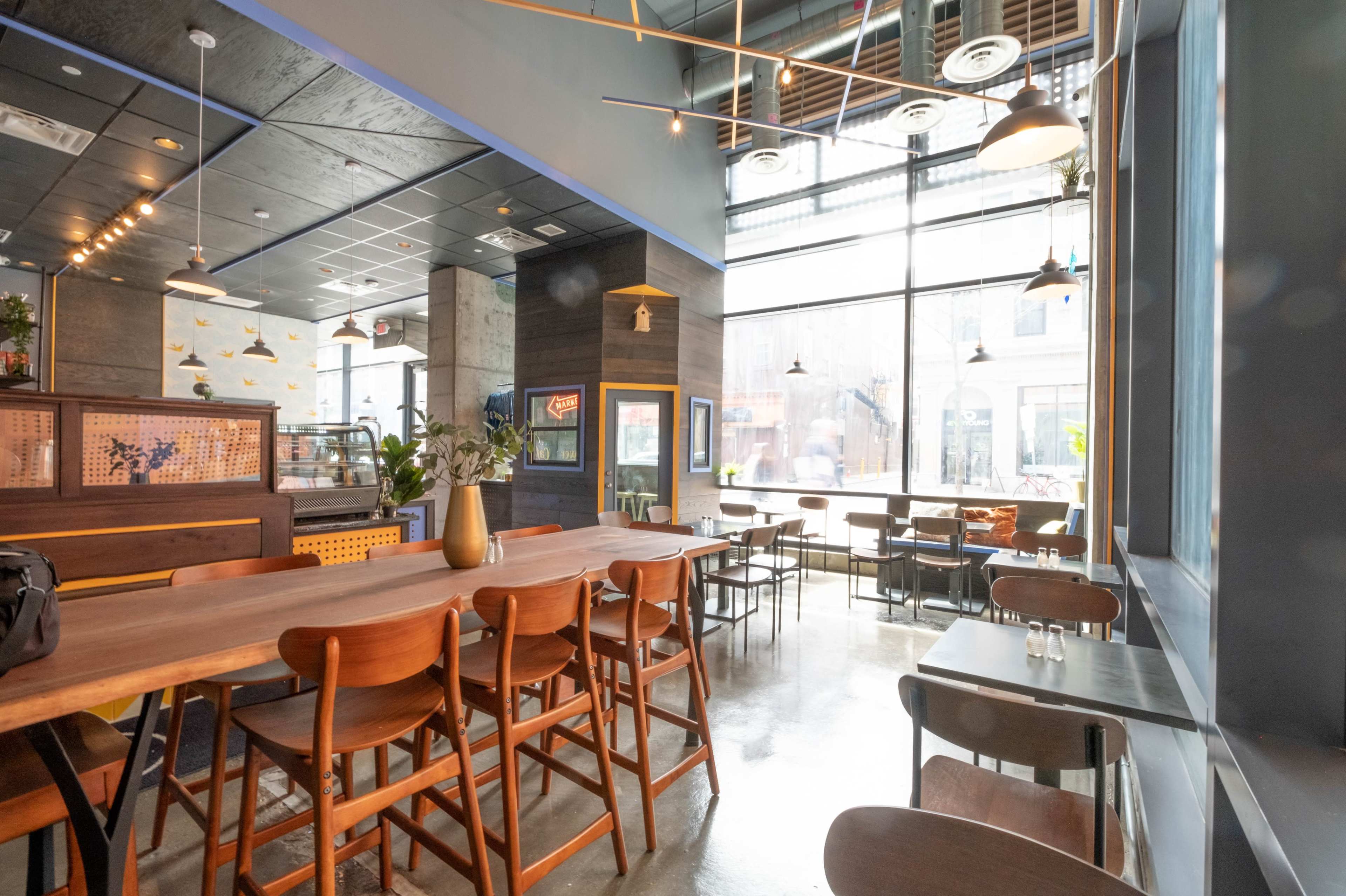 The image shows a modern restaurant interior with wooden tables, high-backed chairs, and large windows allowing natural light to enter.
