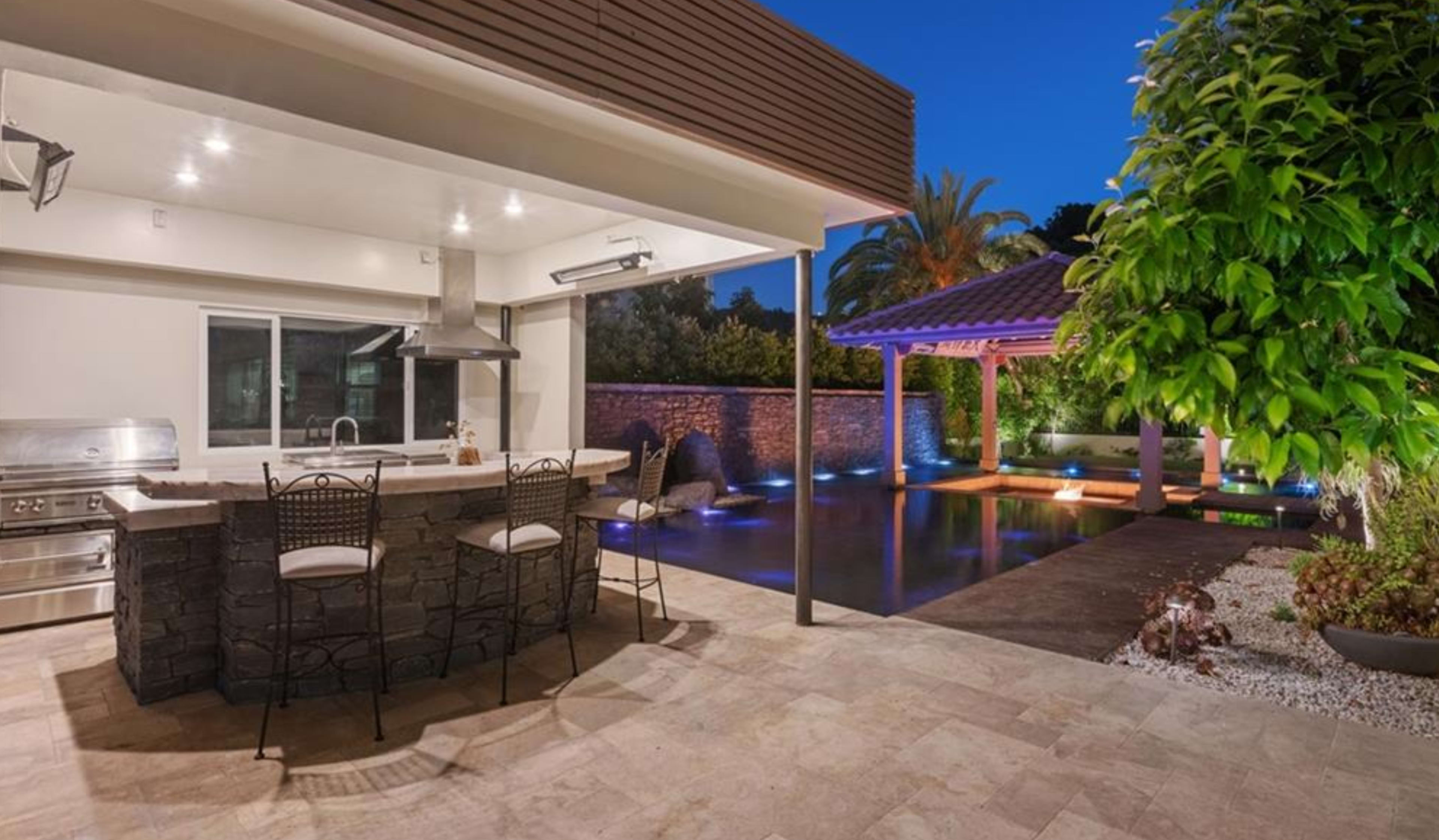 The image shows a backyard with a modern outdoor kitchen featuring a bar, a swimming pool, and a covered seating area illuminated by soft lighting.