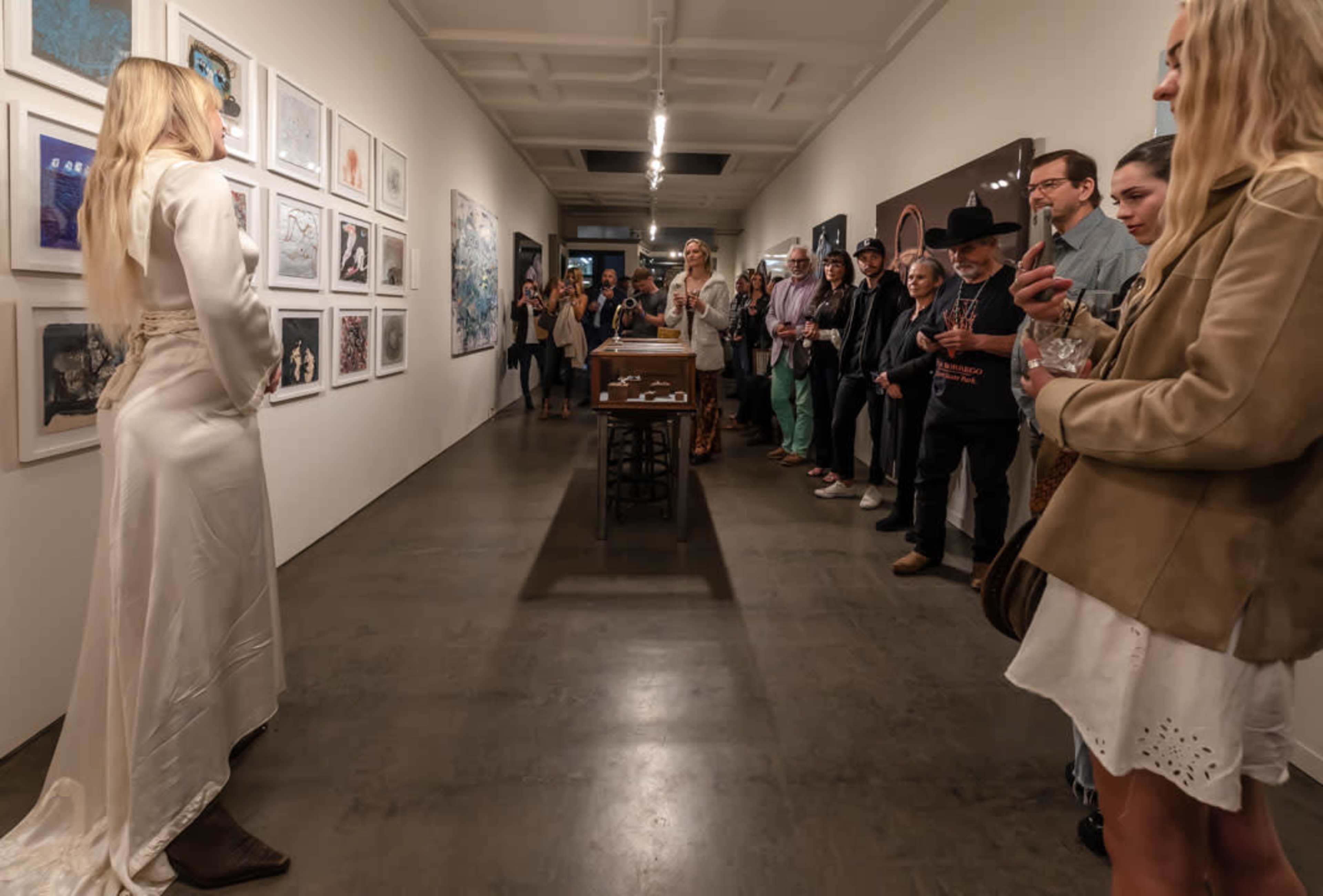 A group of people watches as a woman in a long white dress stands in front of an art display in a gallery.