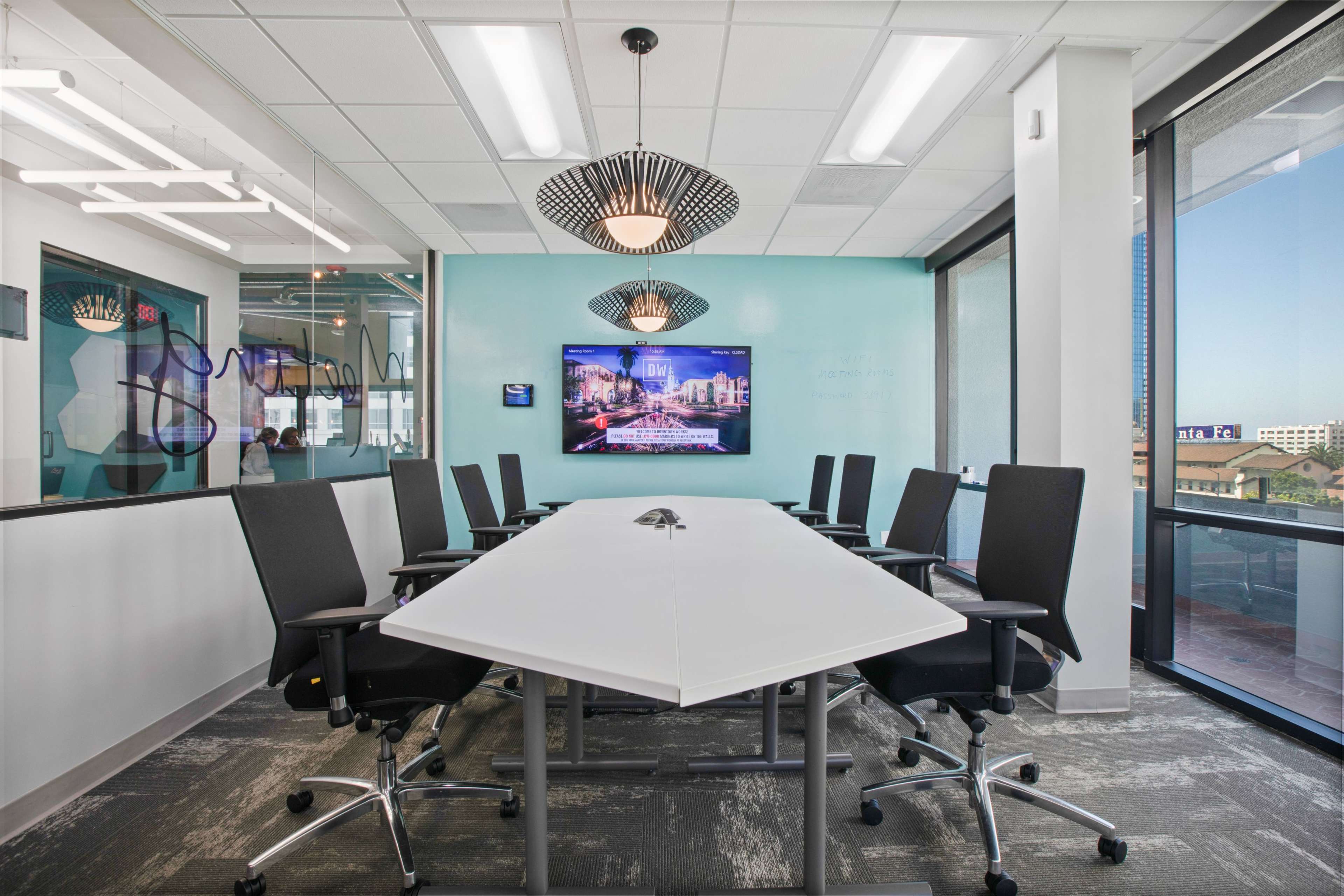 A modern conference room features a hexagonal white table, ergonomic black chairs, large windows, and a wall-mounted TV displaying a cityscape.