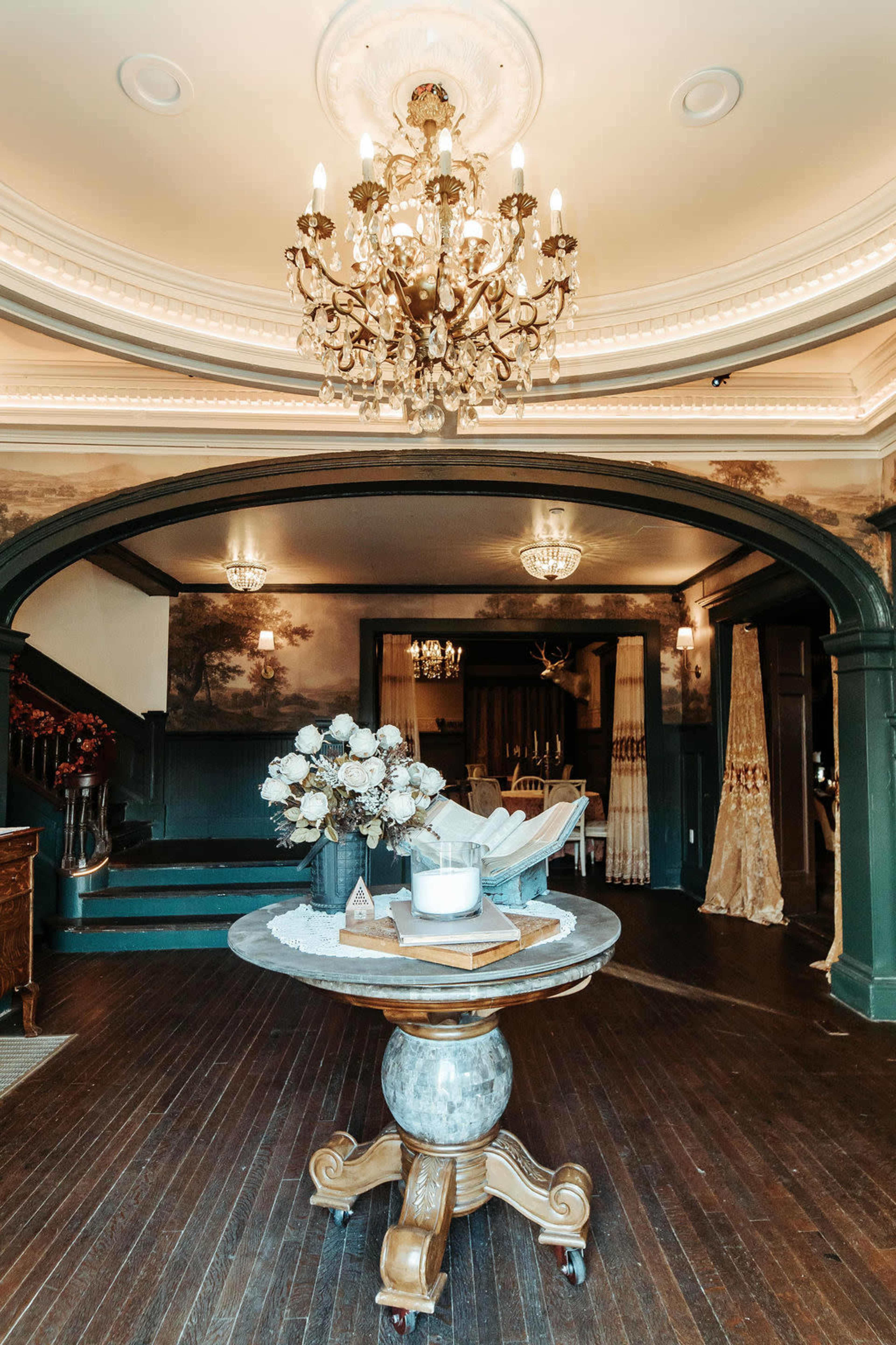 The image shows an elegantly designed foyer featuring a large chandelier, a round table with floral arrangements, and a staircase leading to another level.