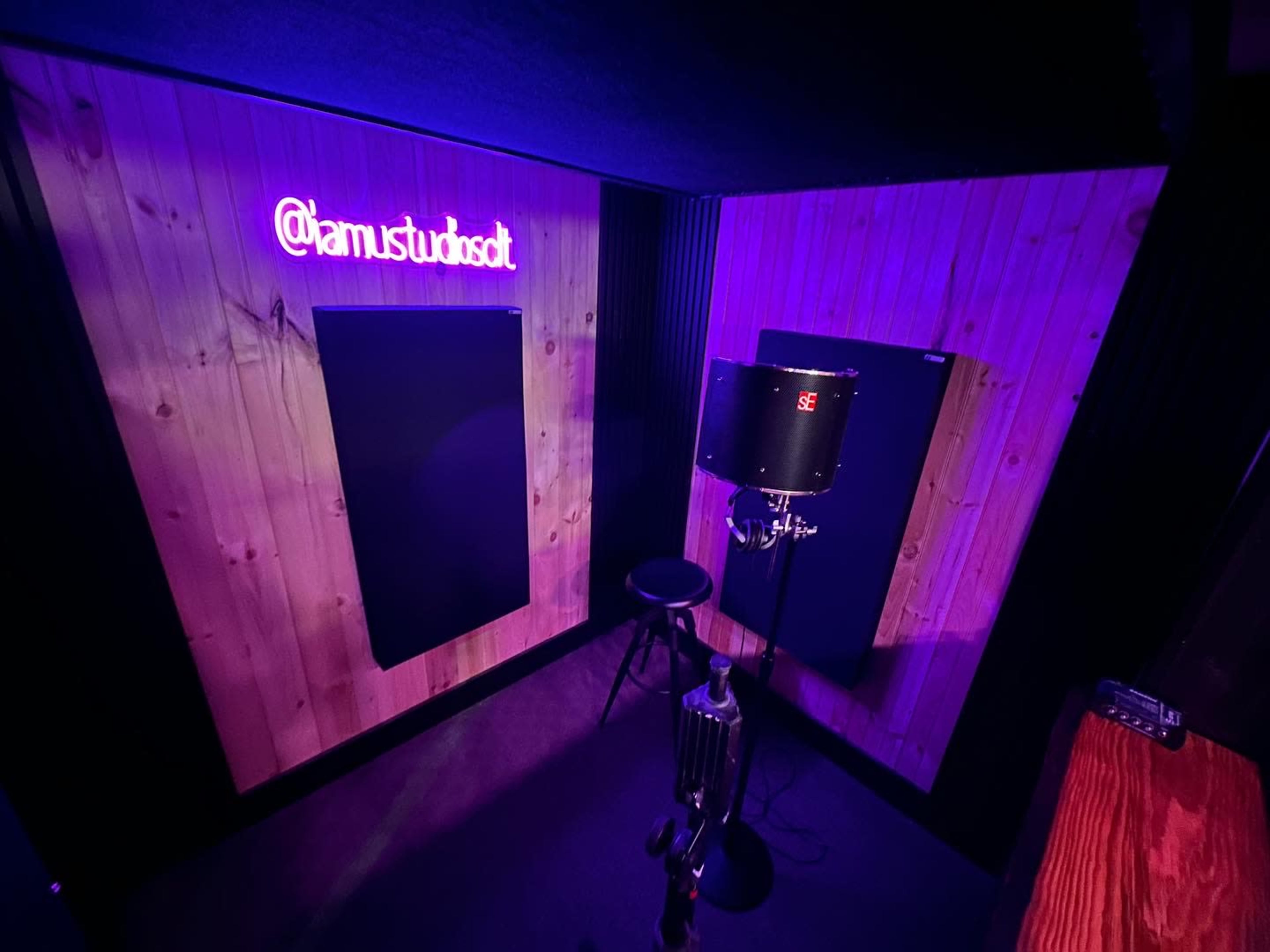 A small soundproof recording booth with wooden paneling, a microphone on a stand, and purple neon lighting displaying the handle @iamstudiosdt.