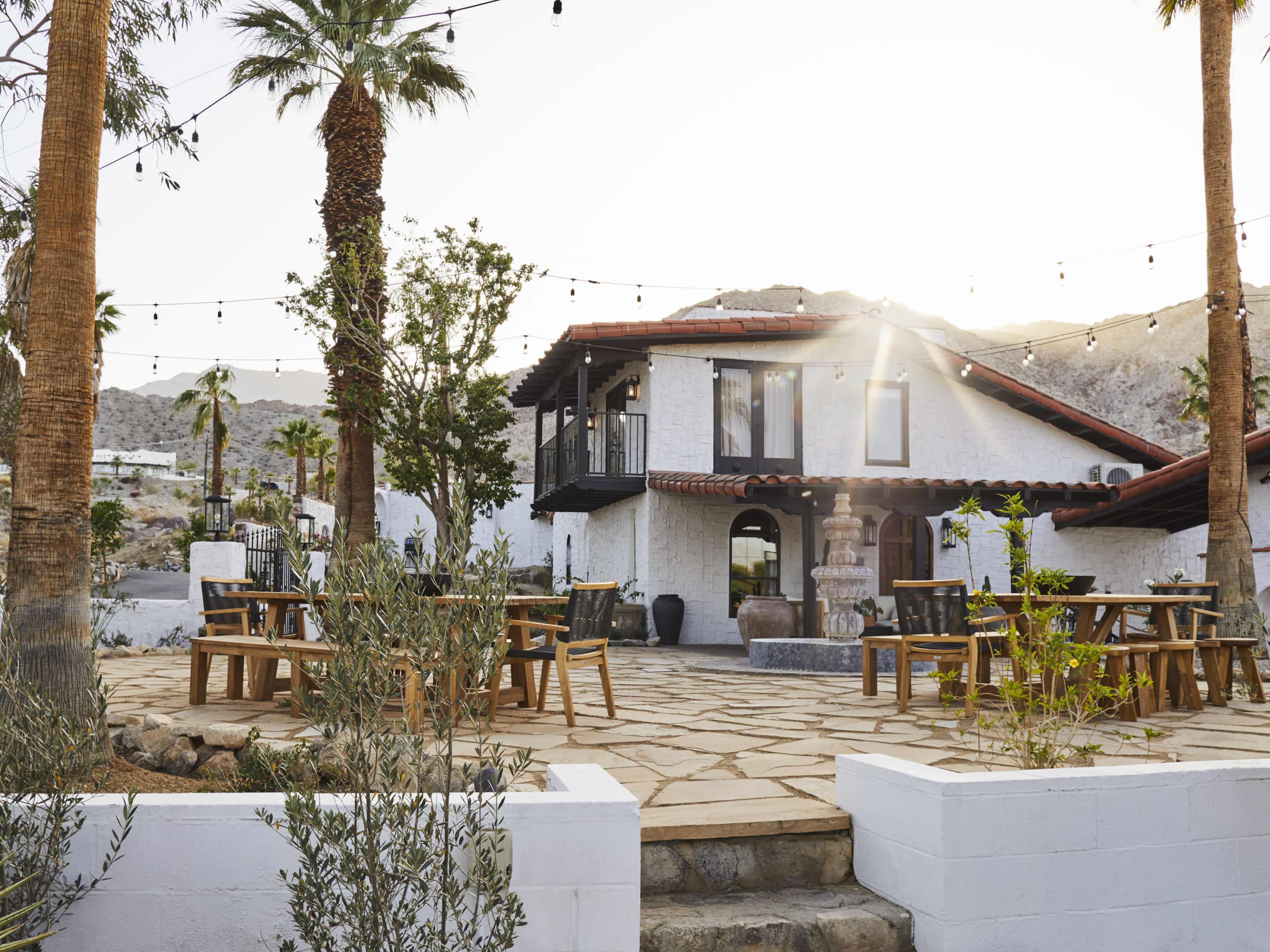 The image shows a modern, outdoor patio area with wooden tables and chairs, surrounded by palm trees and a white building in the background, under a clear sky.