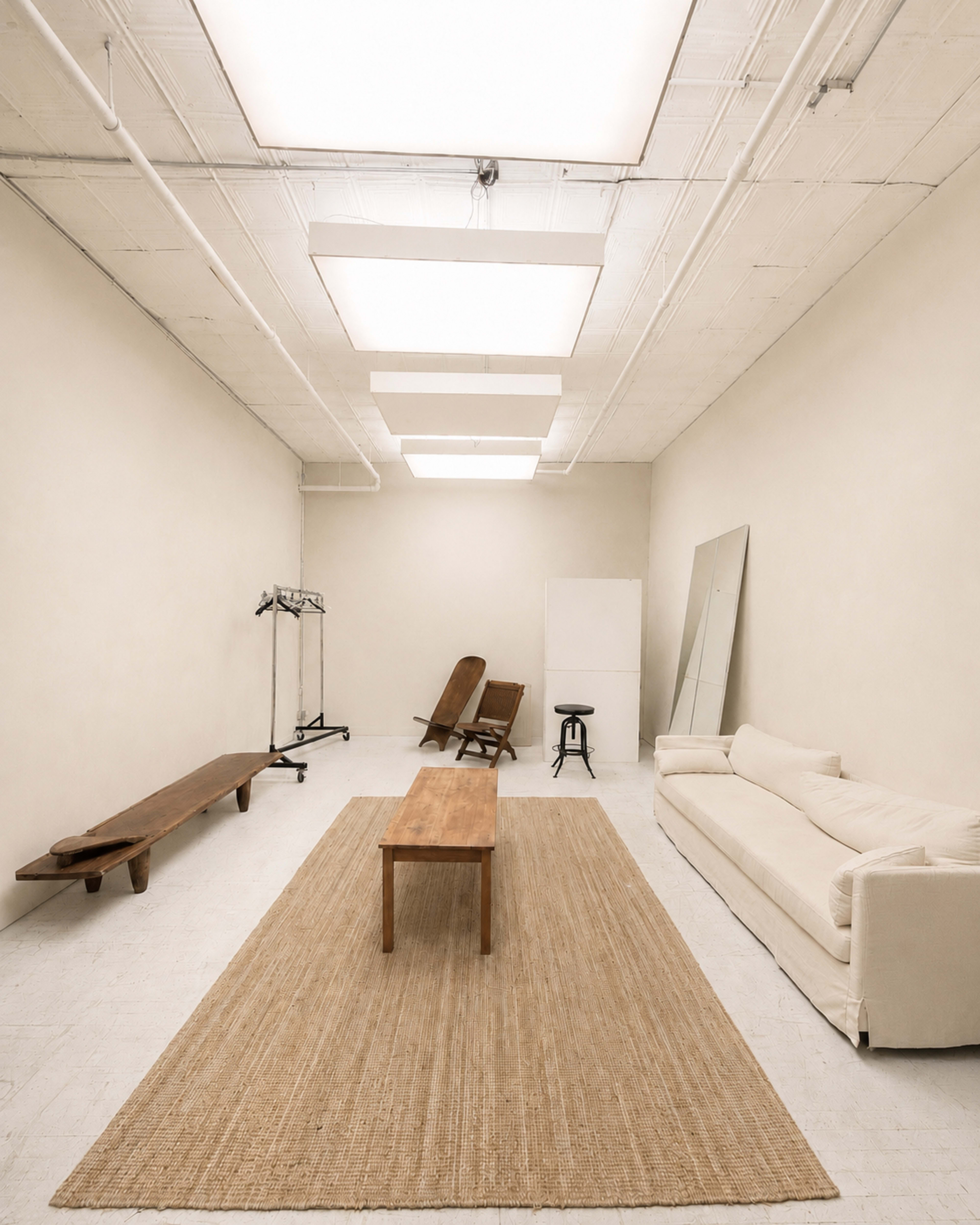 The image depicts a spacious, minimalist room with a wooden table, a light-colored sofa, and a large rug, illuminated by overhead skylights.