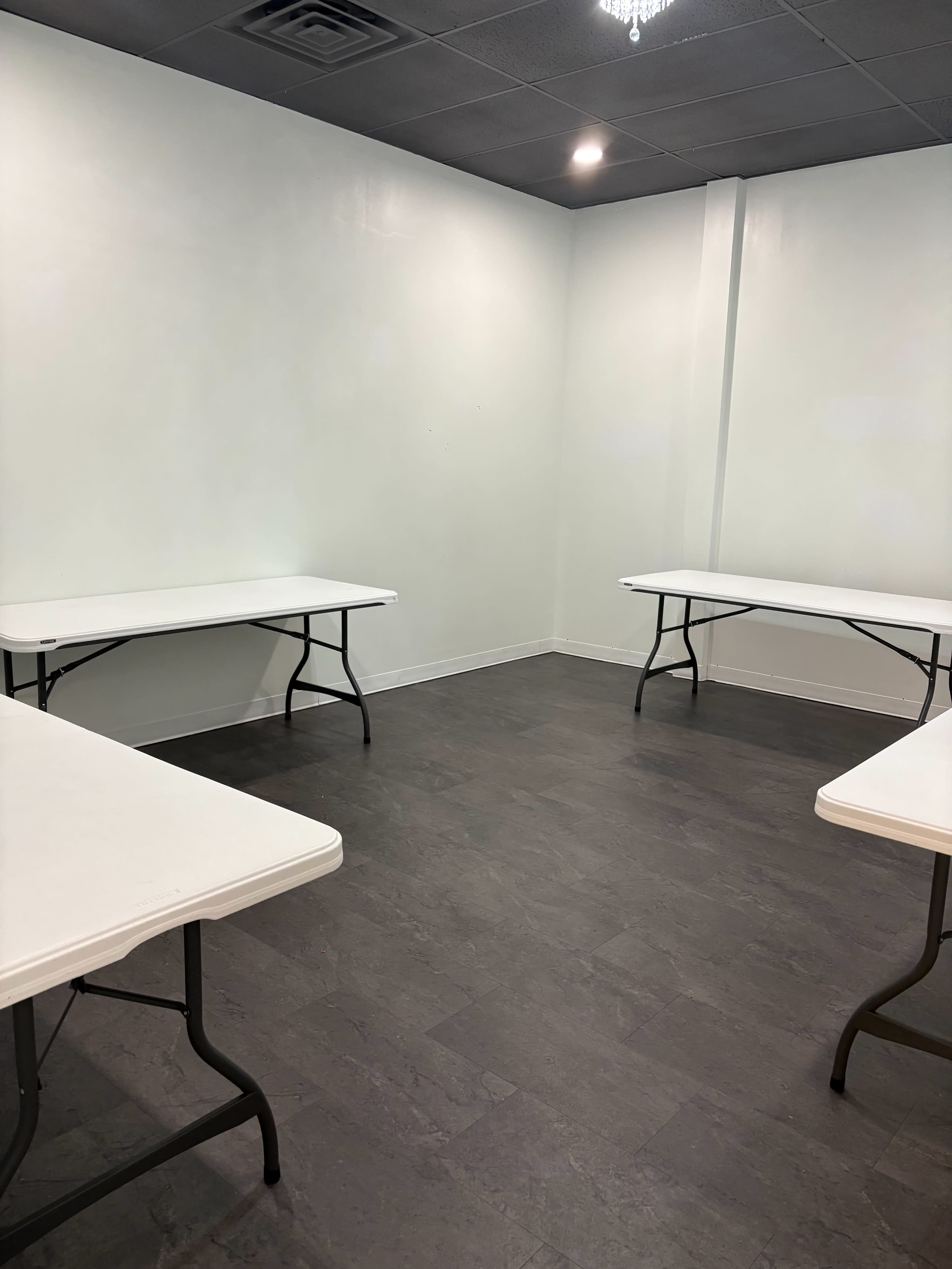 The image shows a sparsely furnished room with four folding tables arranged in a square pattern on a dark floor.
