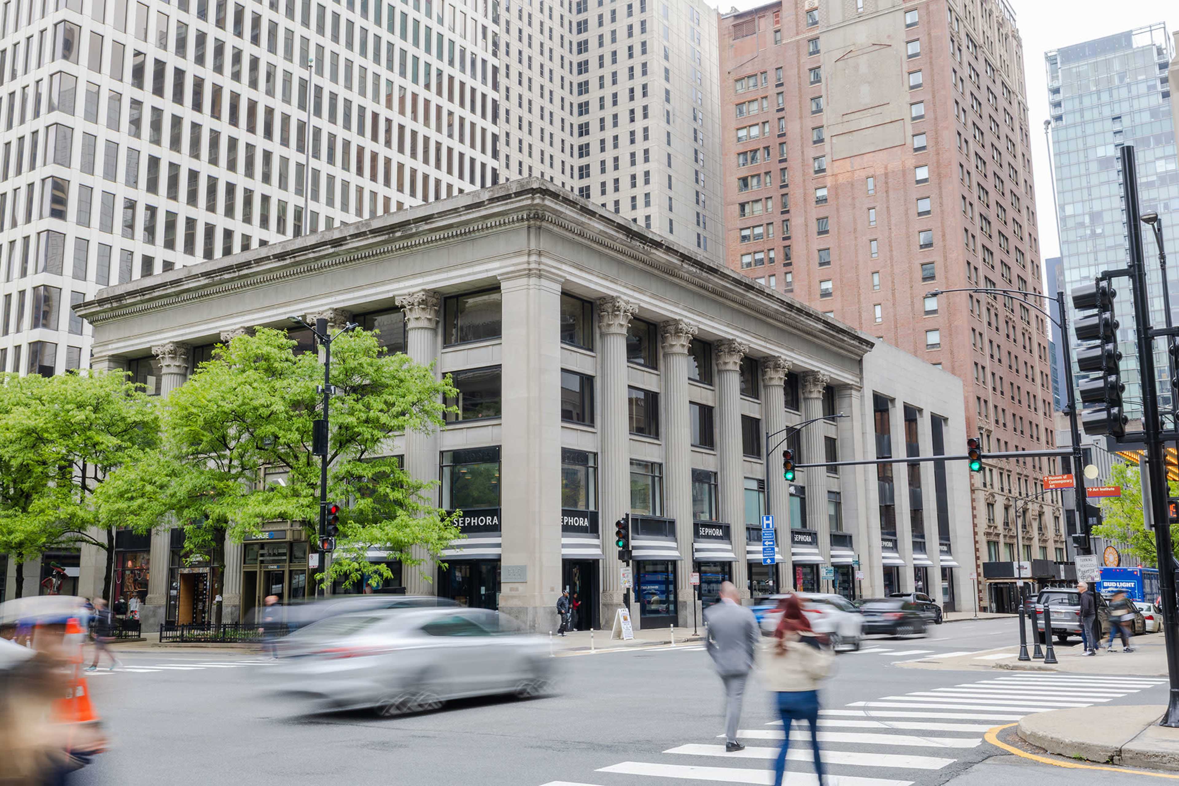 A busy urban intersection features a historic building with tall columns, flanked by modern skyscrapers, as cars and pedestrians navigate the crosswalk.
