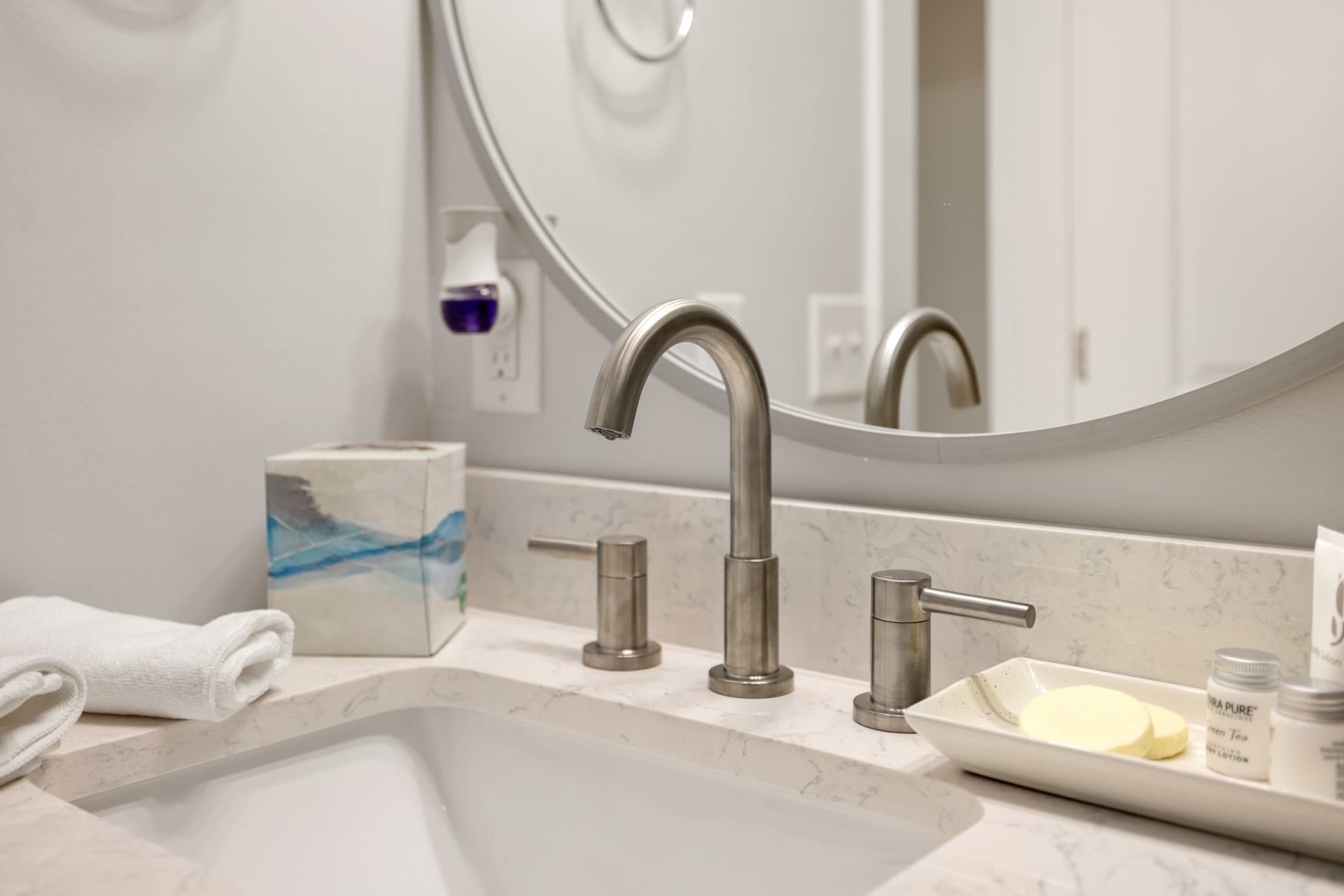 A modern bathroom sink with a circular mirror, featuring a brushed metal faucet and a small tray containing skincare products.