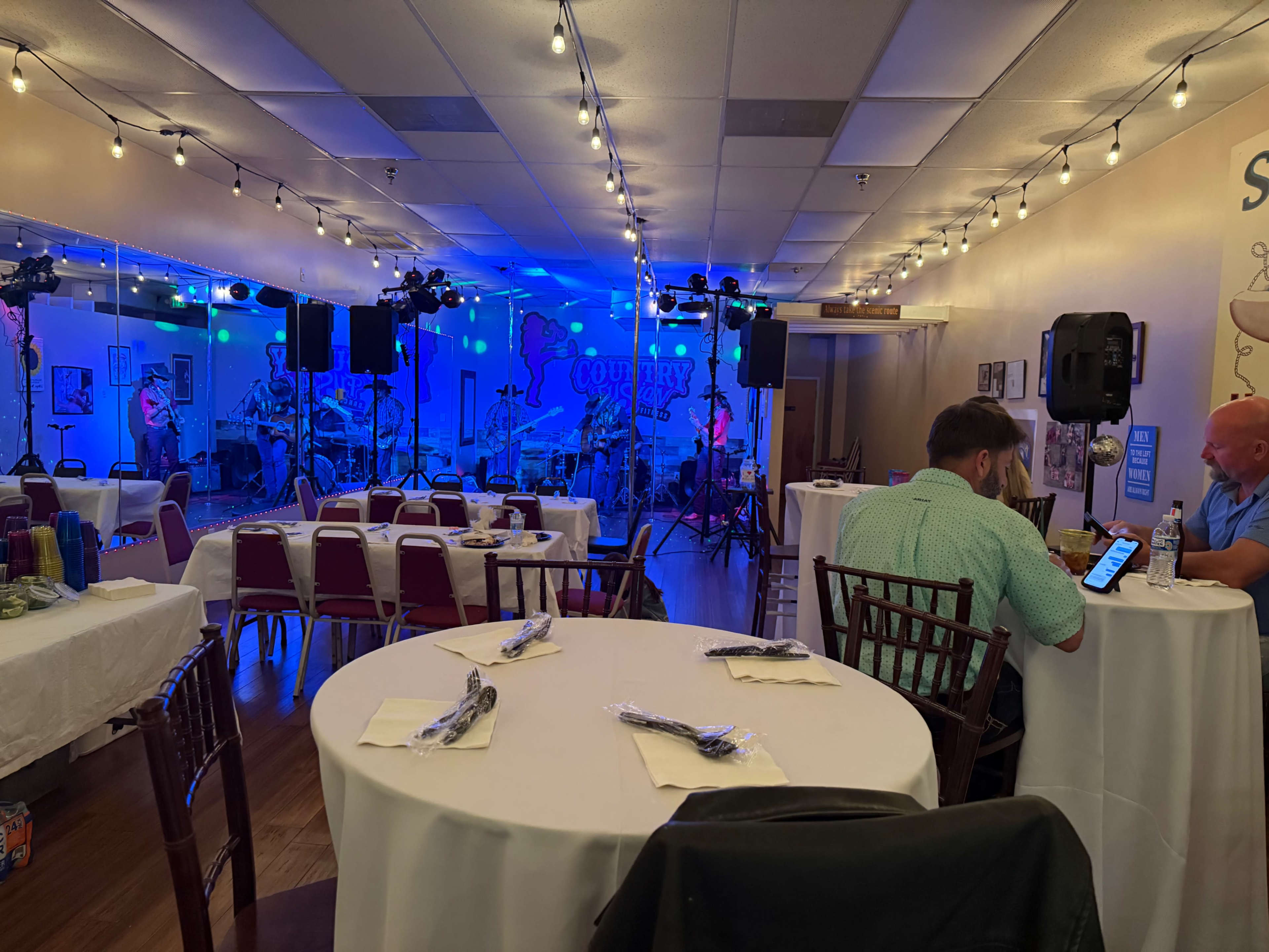 The image shows a spacious live music venue with a stage for a band, tables set for dining, and colorful lights illuminating the space.