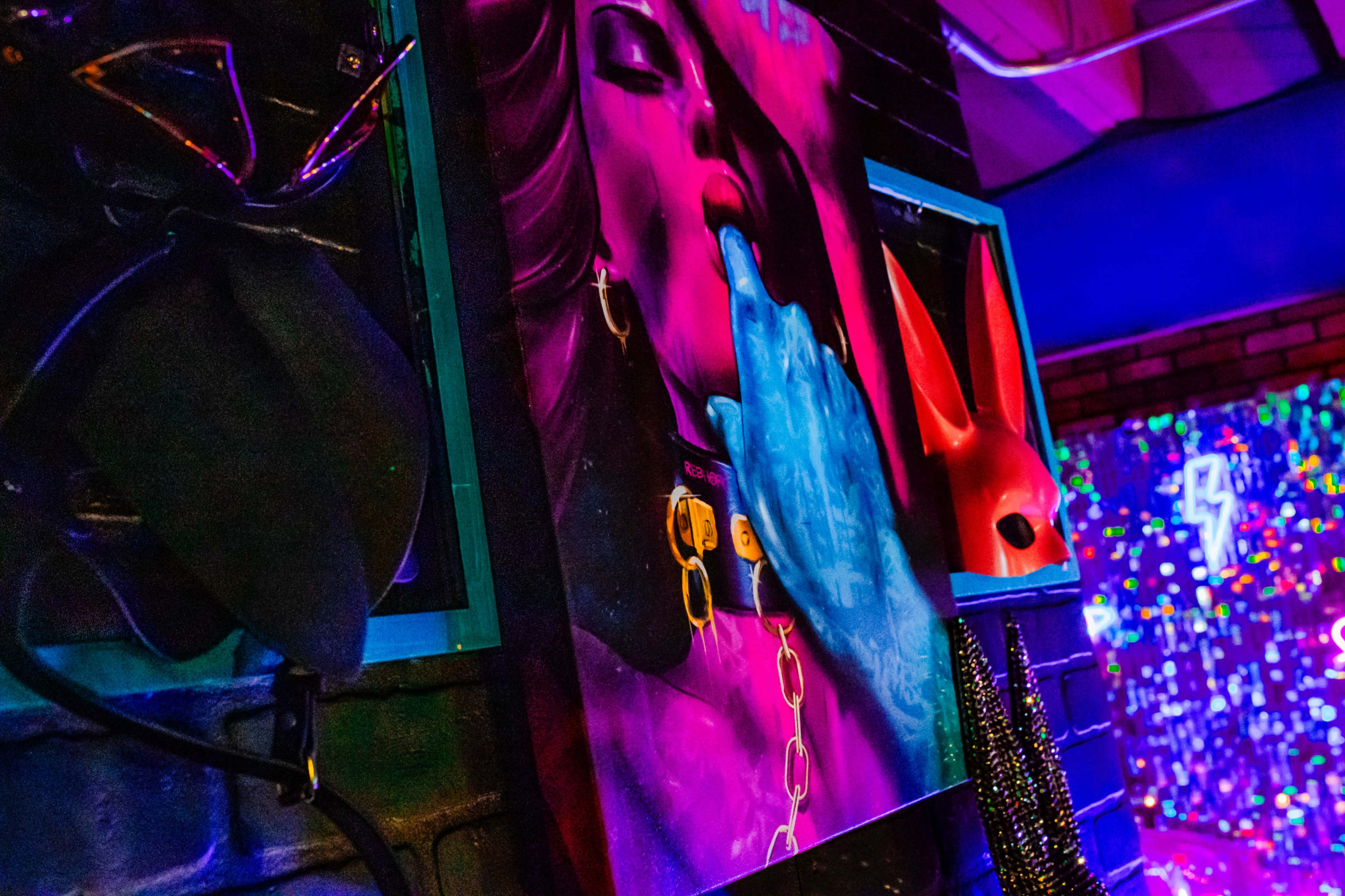 The image features a colorful wall adorned with framed artwork depicting a figure with a striking pose, alongside various accessories including masks and chains.