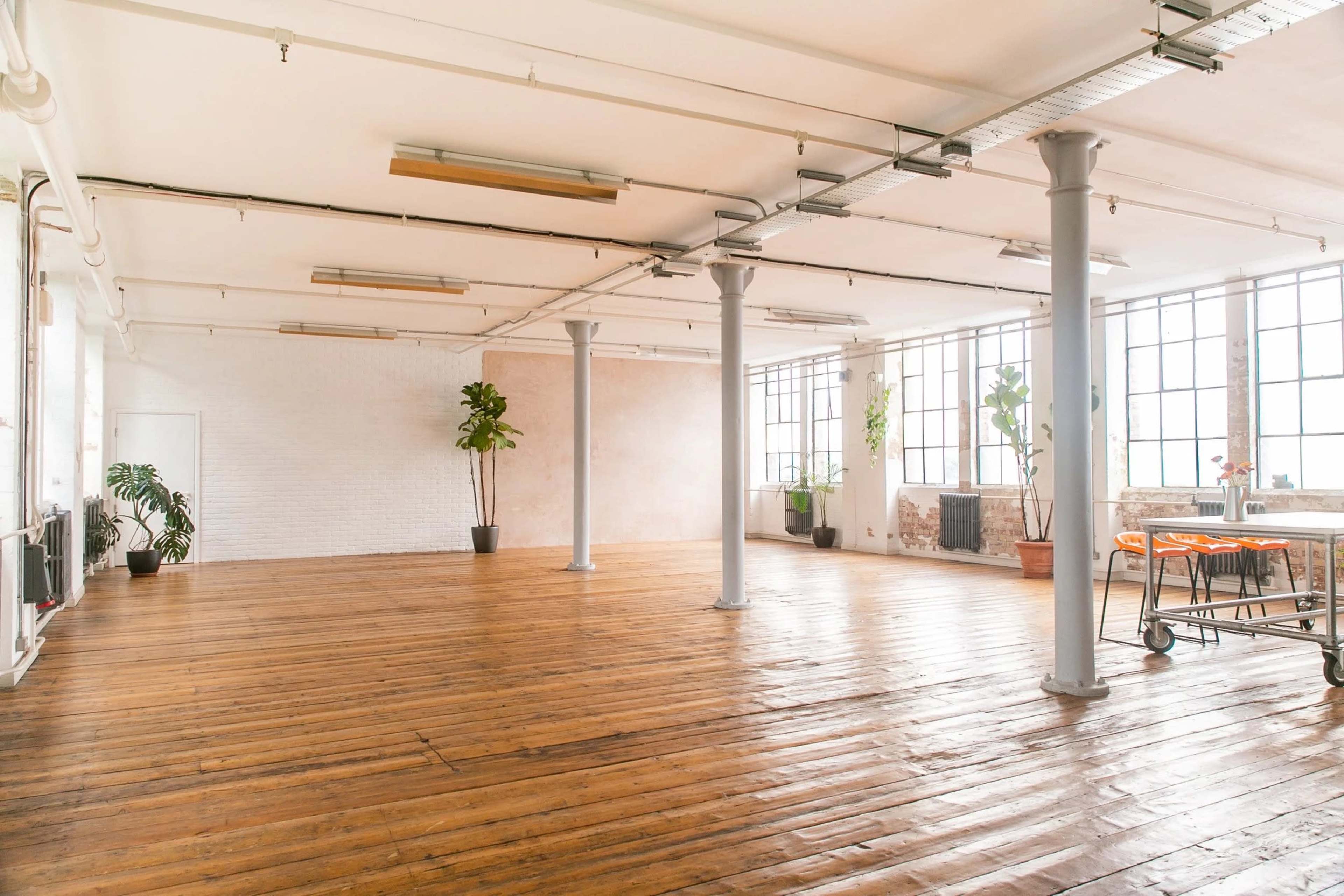 The image shows a spacious, empty interior of a room with wooden floors, large windows, and a few plants in pots.