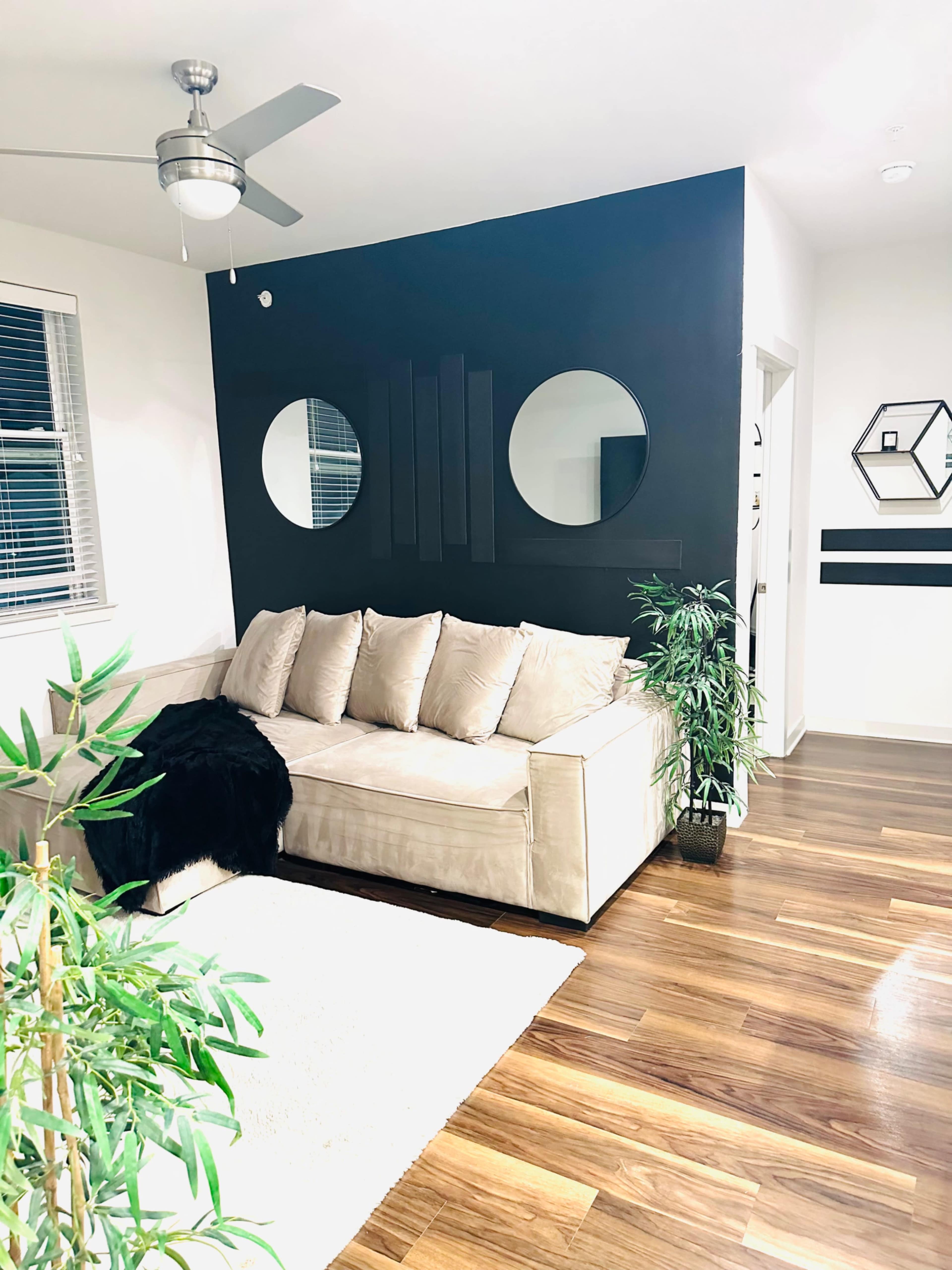 The image shows a modern living room with a beige sofa, a black accent wall featuring circular mirrors, and a plant beside a white rug on hardwood flooring.