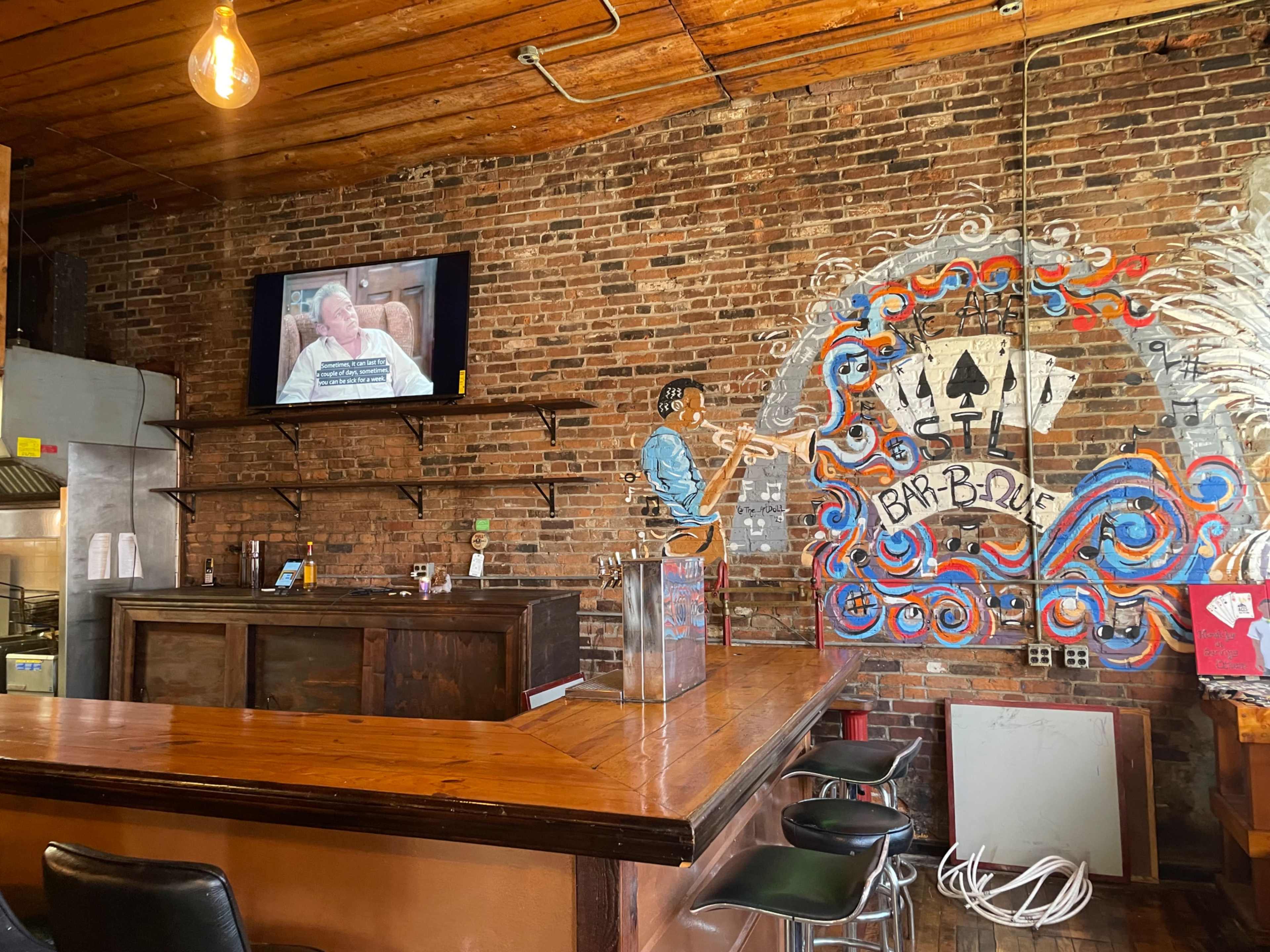The interior of a bar-b-que restaurant features a wooden bar with a television displaying a program, against a backdrop of exposed brick walls and colorful artwork.