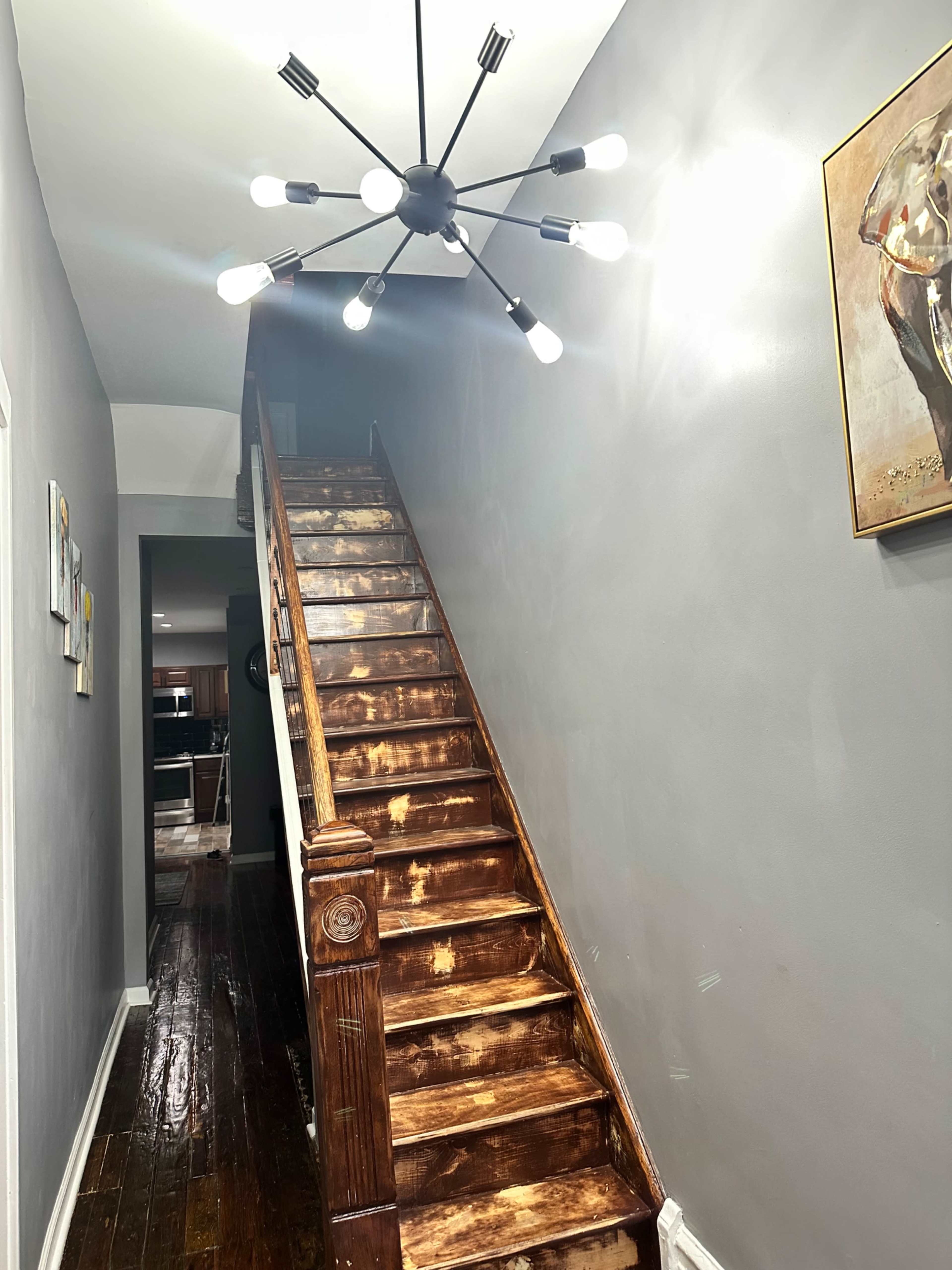The image shows a wooden staircase leading up, illuminated by a modern, starburst-style light fixture against a gray wall.