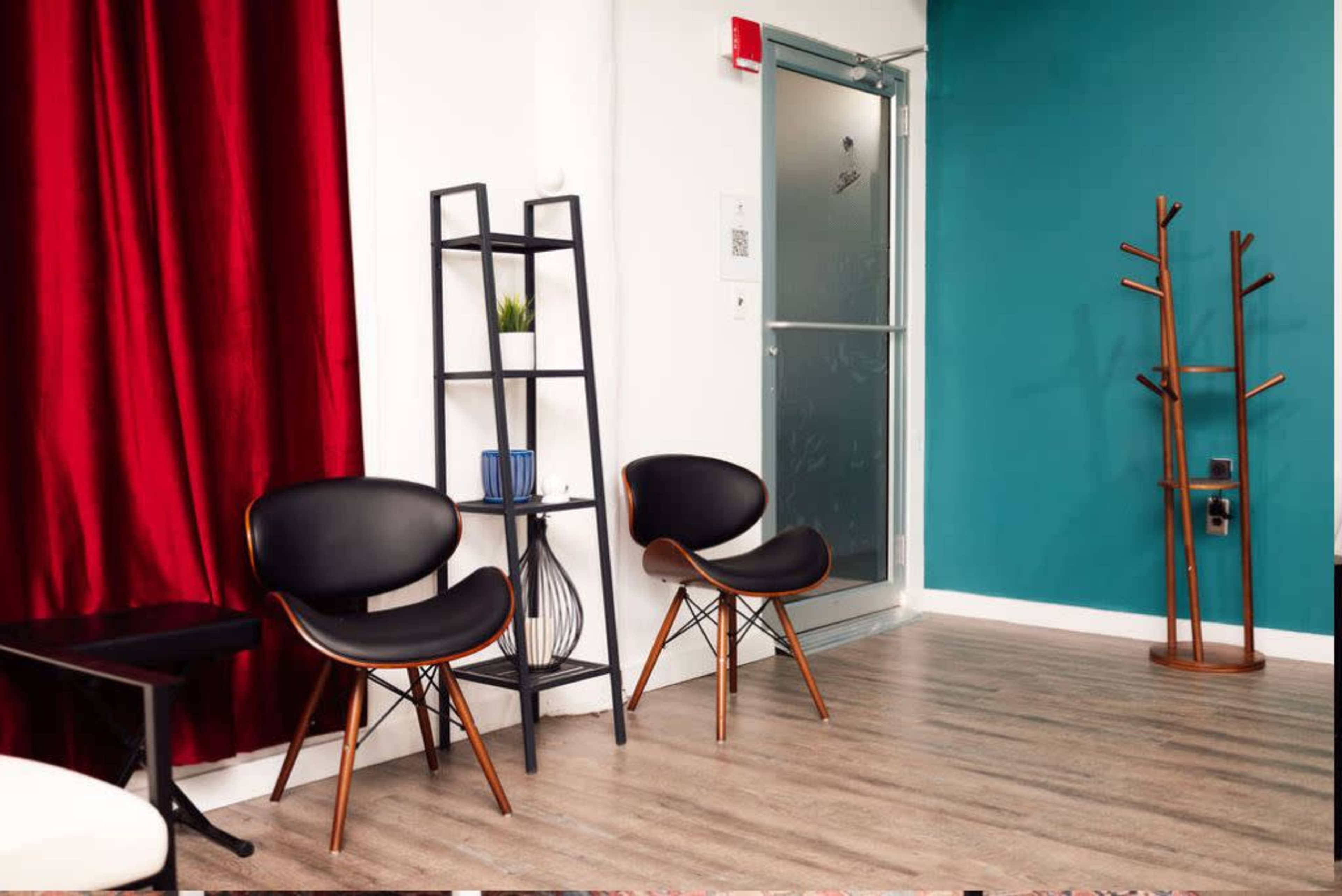 A modern waiting area featuring two black chairs, a tall black shelf with decorative items, a coat rack, and a door against a wall with teal paint and red curtains.