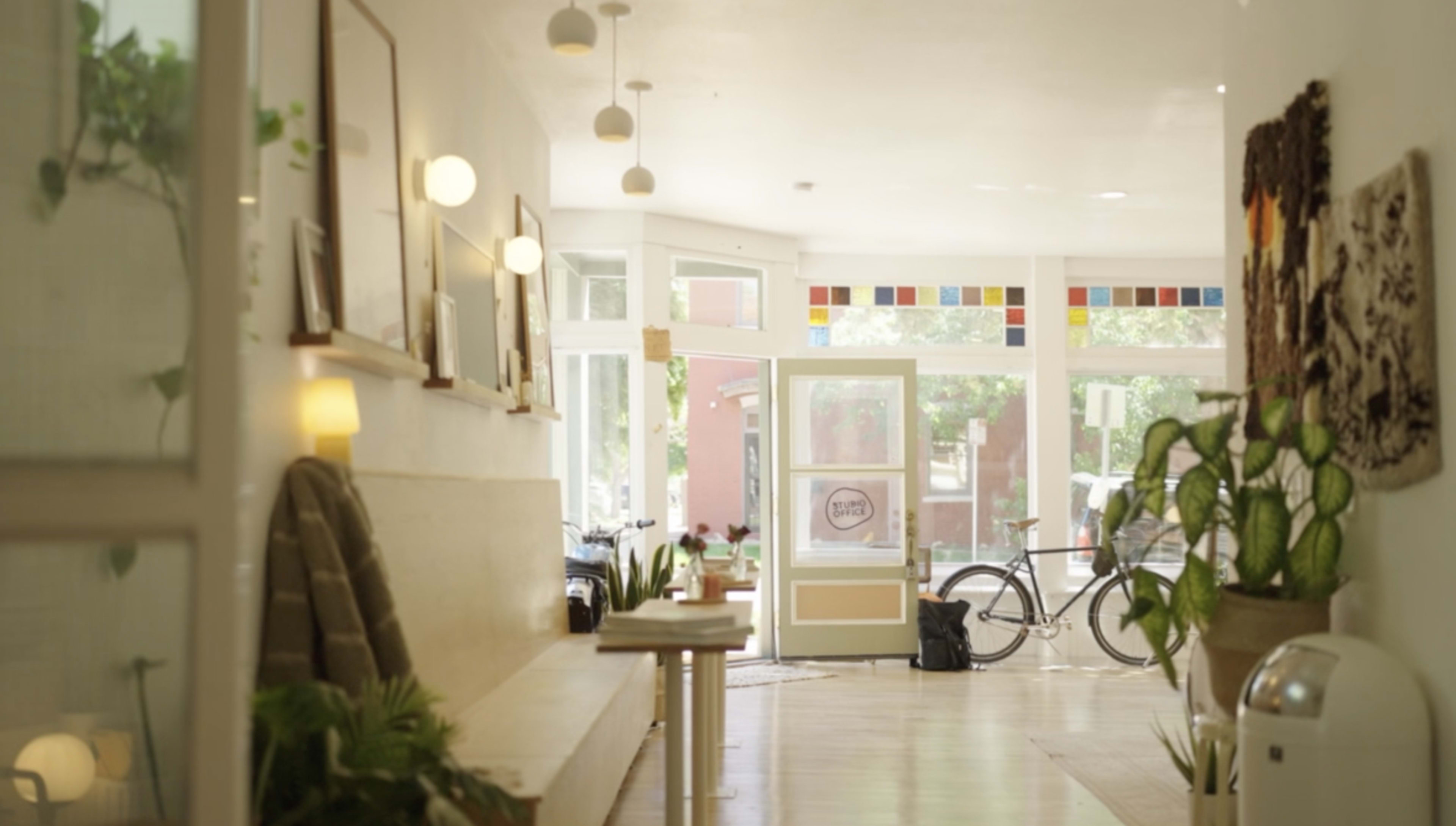 The image shows a bright hallway of a café with a bicycle parked near the entrance and plants adorning the space.