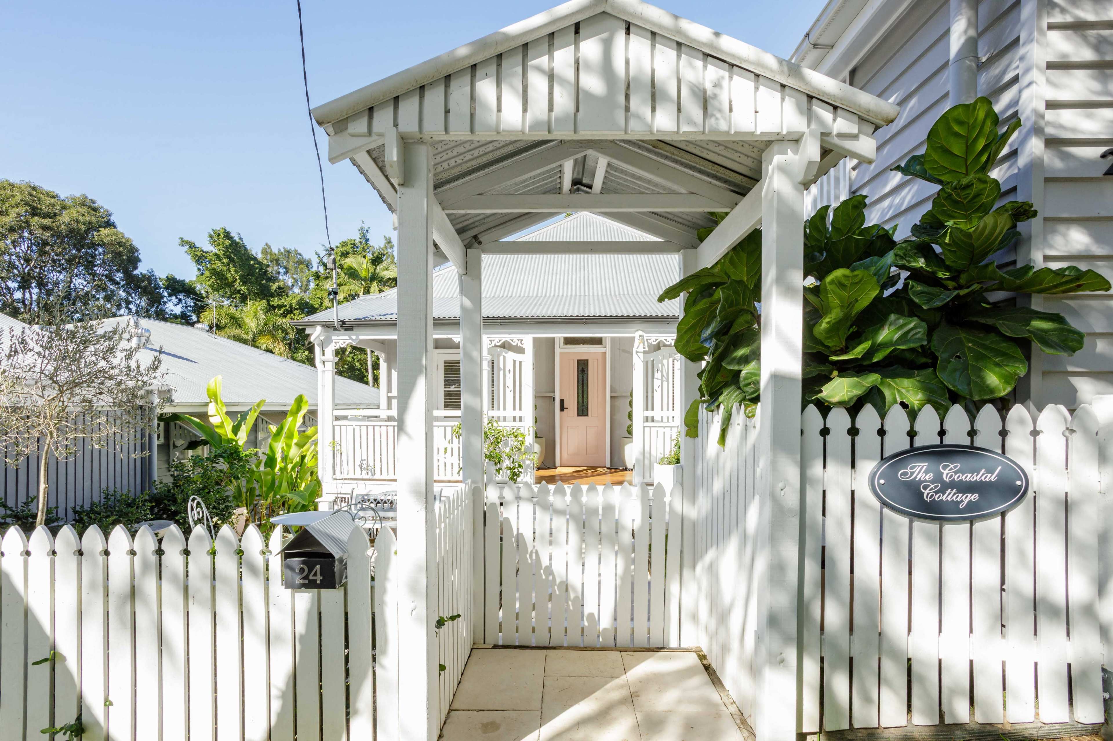 The image shows a white cottage entrance featuring a covered walkway, a picket fence, and lush greenery surrounding the pathway.