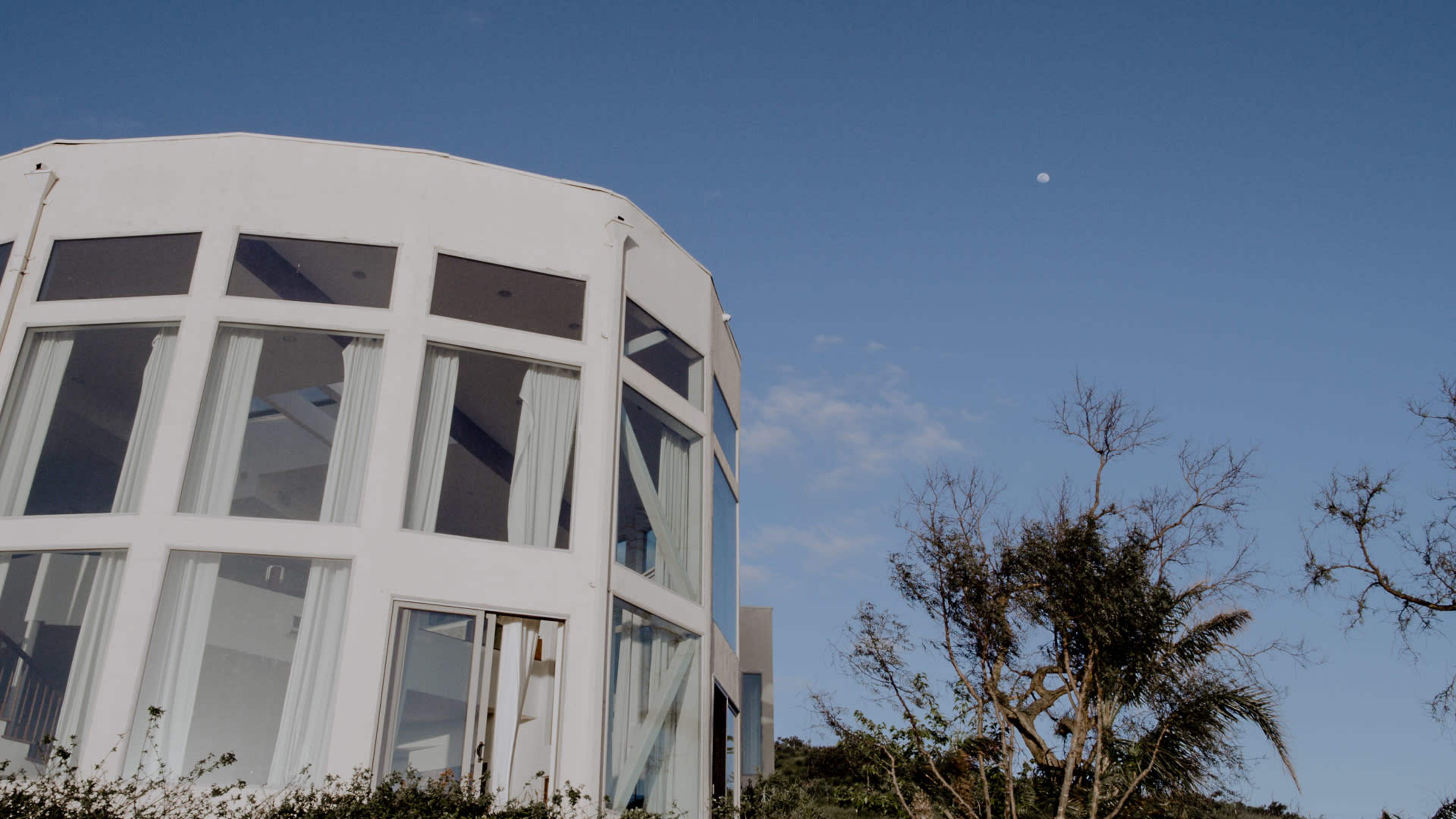 A modern, circular white building with large glass windows is partially visible under a clear blue sky, with a moon and sparse trees in the background.