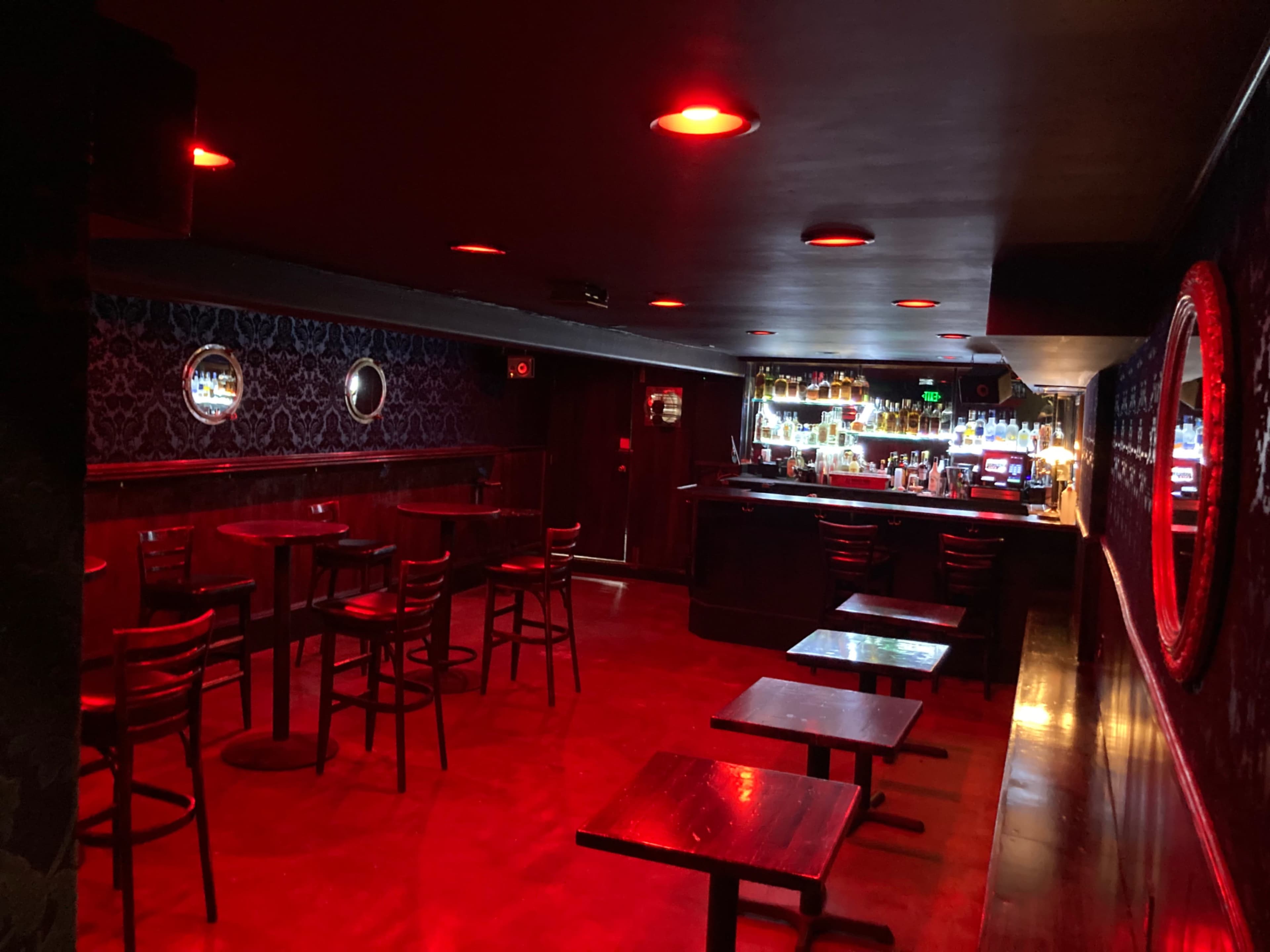 A dimly lit bar features a row of empty tables and a lighted bar counter in the background.