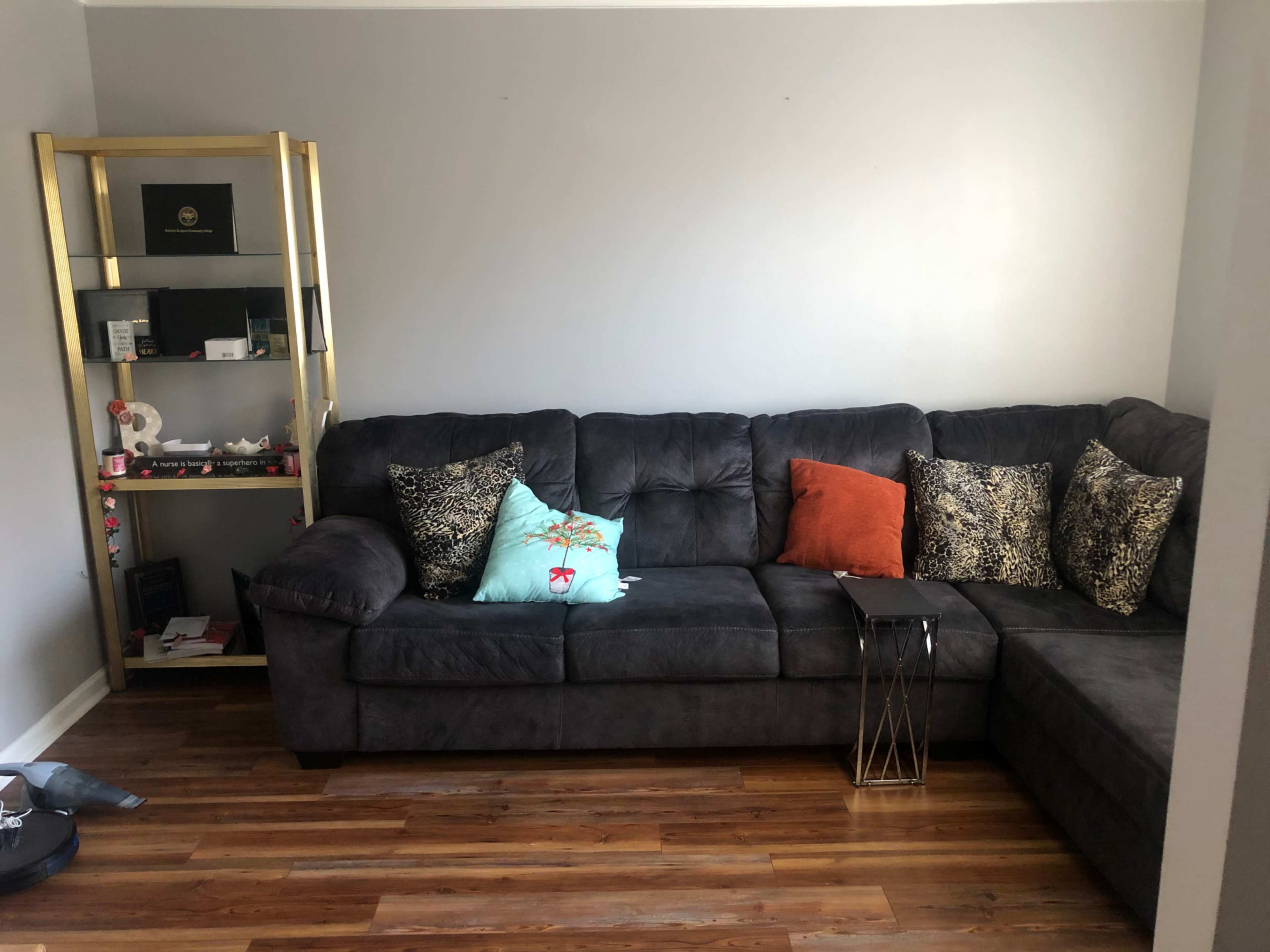 A living room featuring a dark gray sectional sofa with decorative pillows, a small metal side table, and a gold shelf displaying various items.