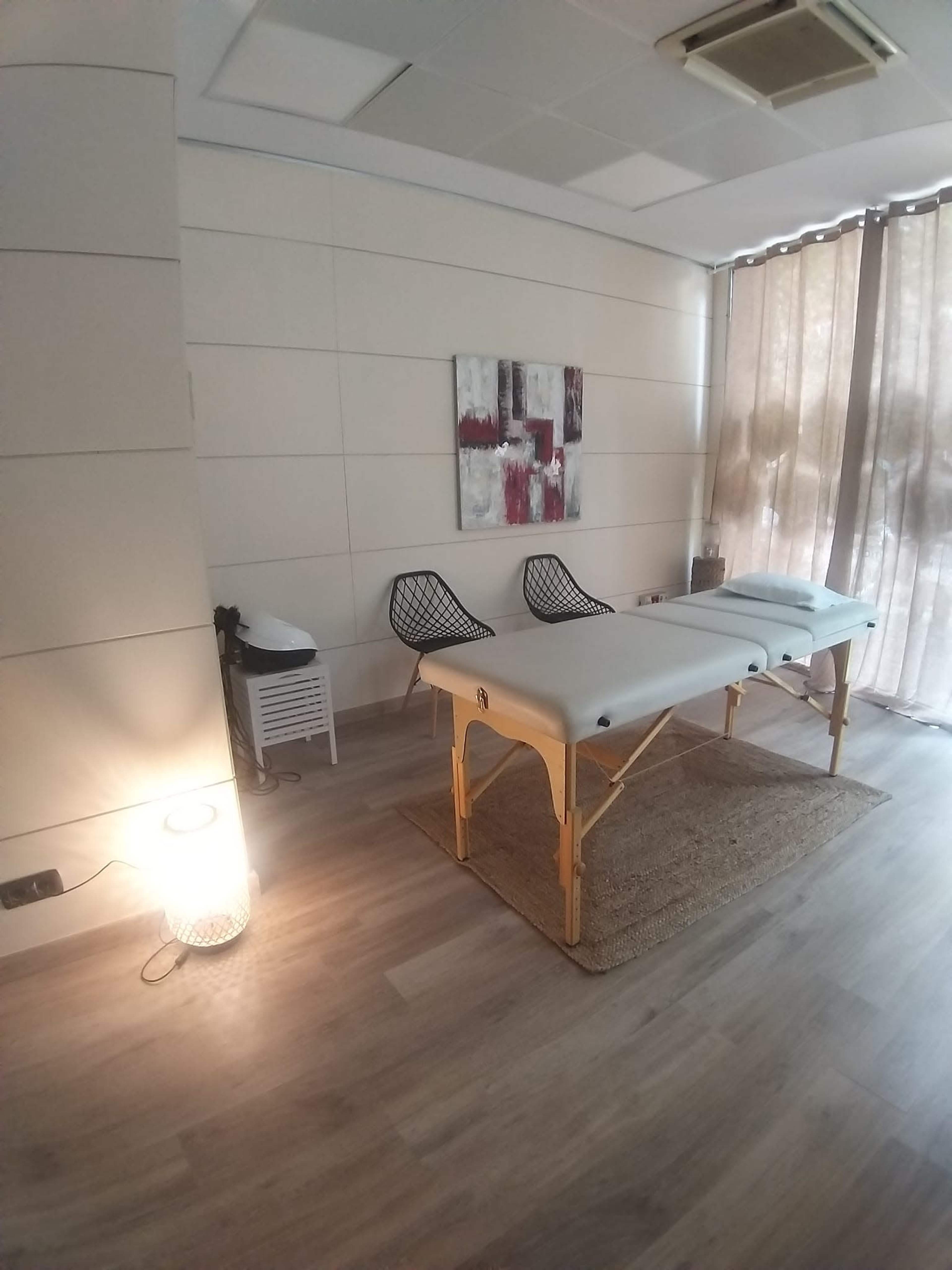 A massage therapy room features a treatment table, two chairs, and a decorative wall art piece, with natural light filtering through sheer curtains.